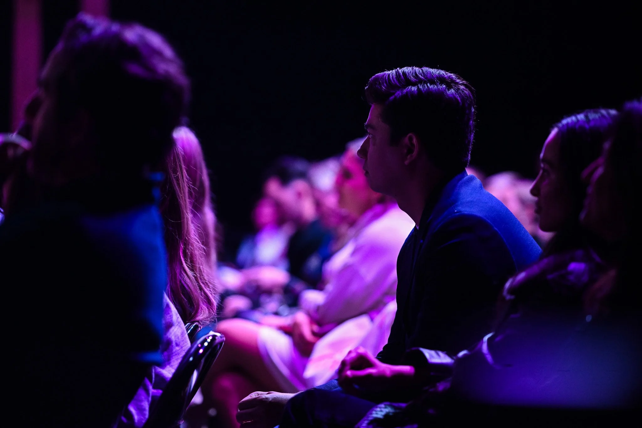 People attending a conference or event, sitting in rows in a dimly lit room with purple lighting.