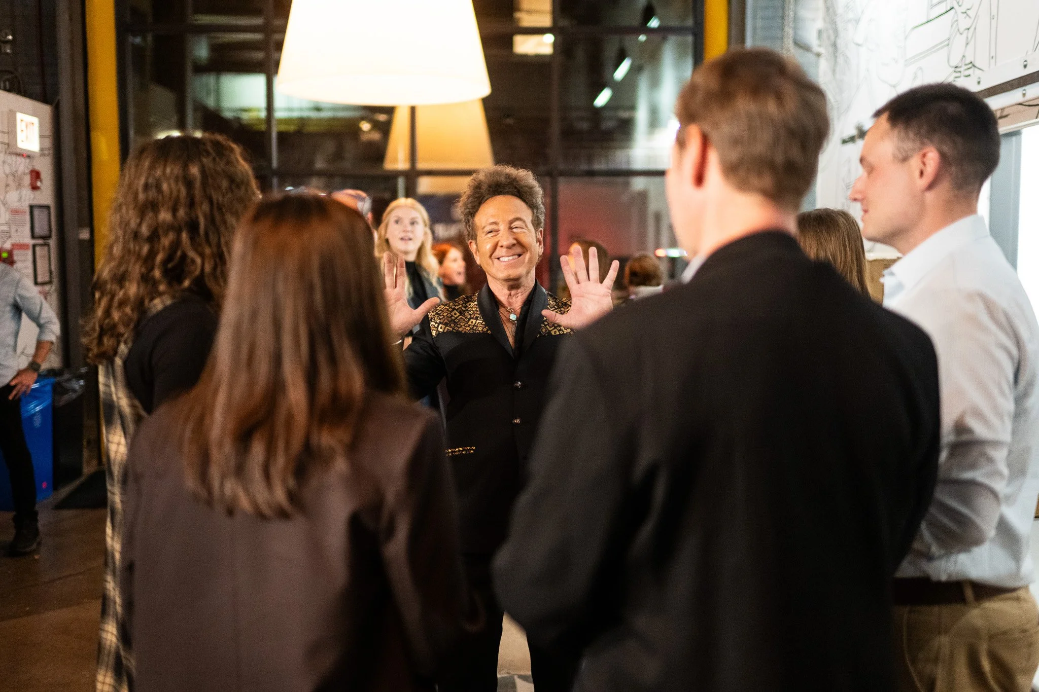 Person smiling and gesturing with hands while speaking to a group of people in an indoor setting.