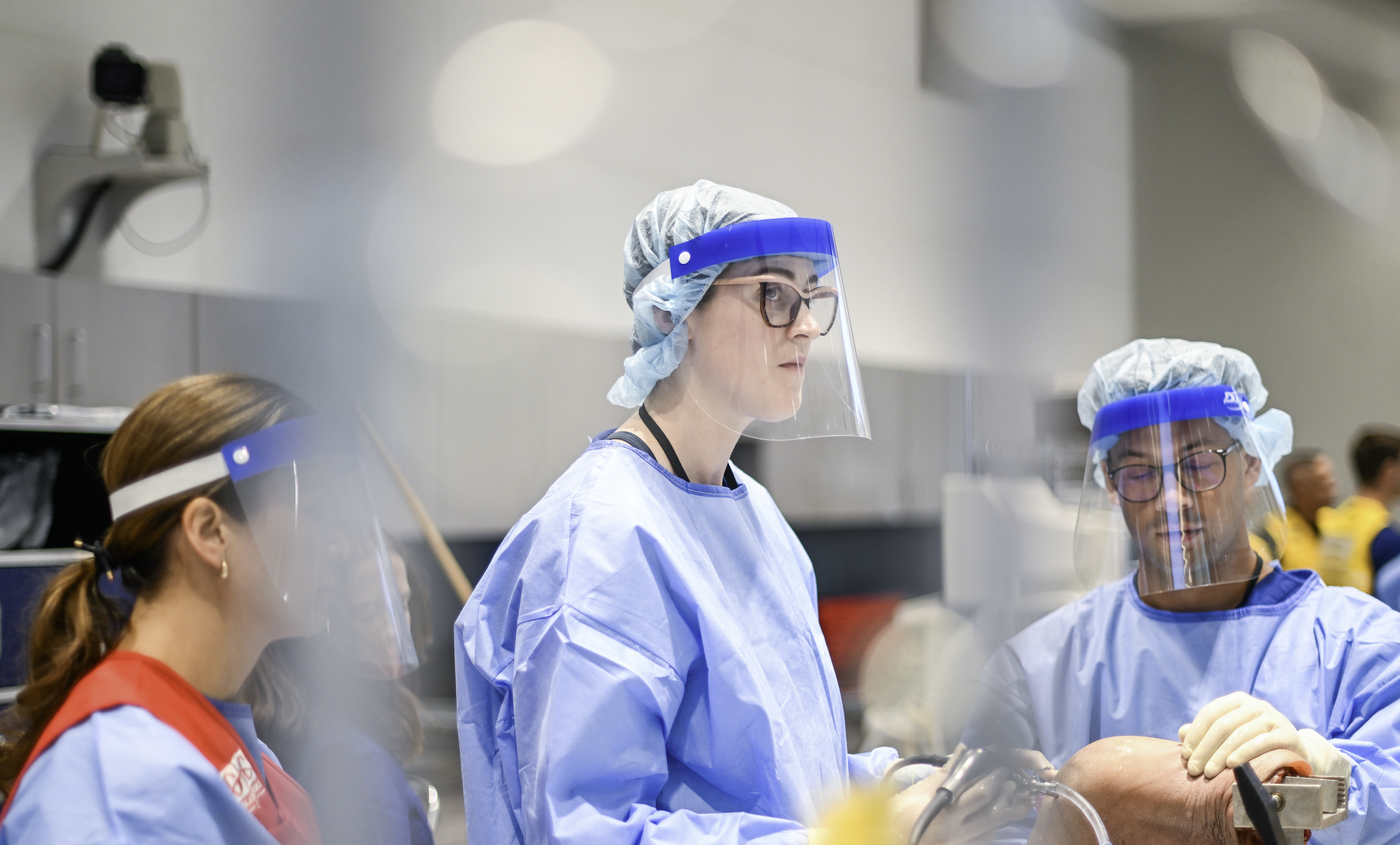 Three healthcare professionals in scrubs and PPE performing a medical procedure in a hospital setting.
