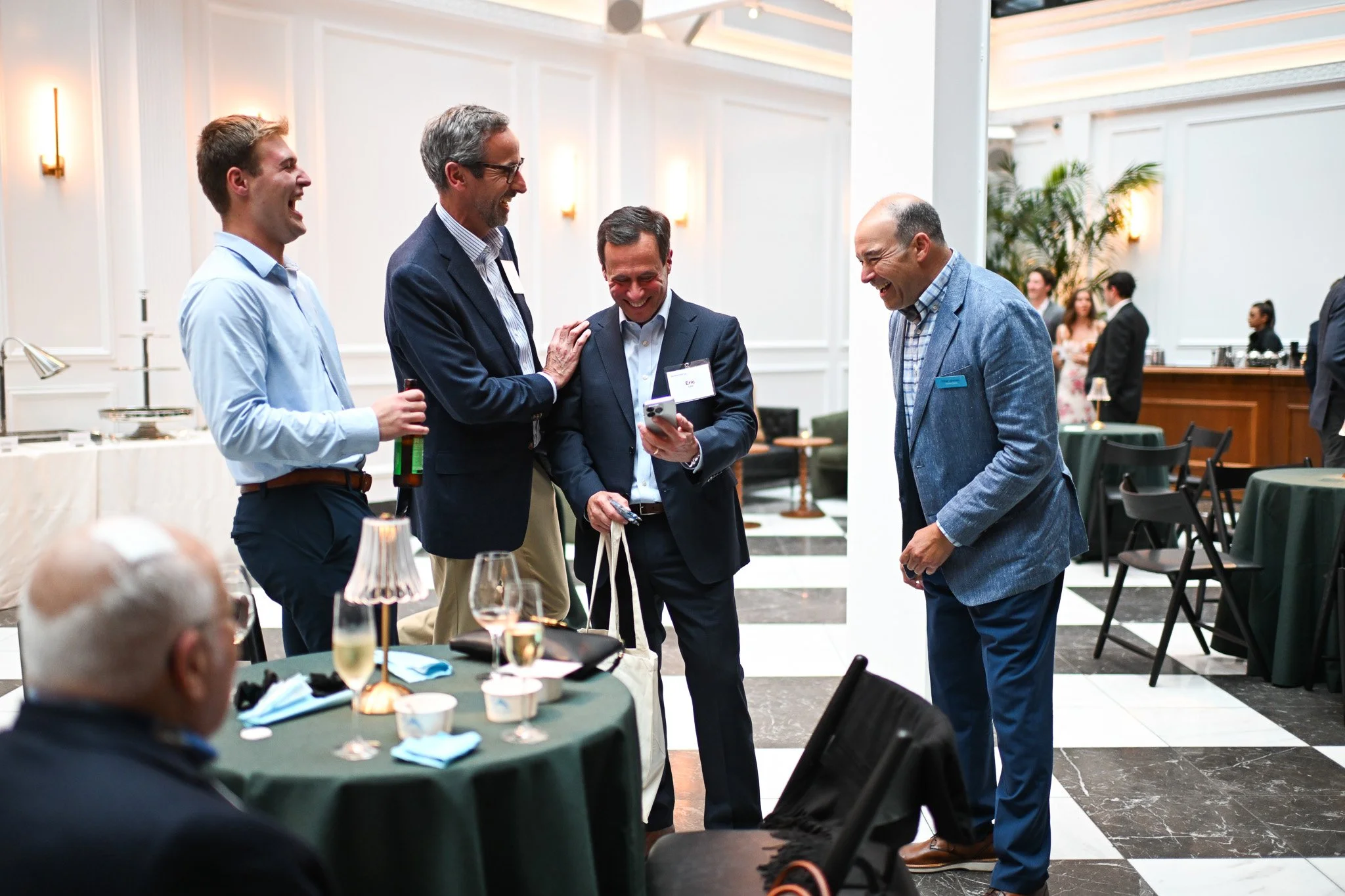 Four men in business attire are standing around a table, laughing and looking at a phone during a networking event or conference in an elegant indoor venue.