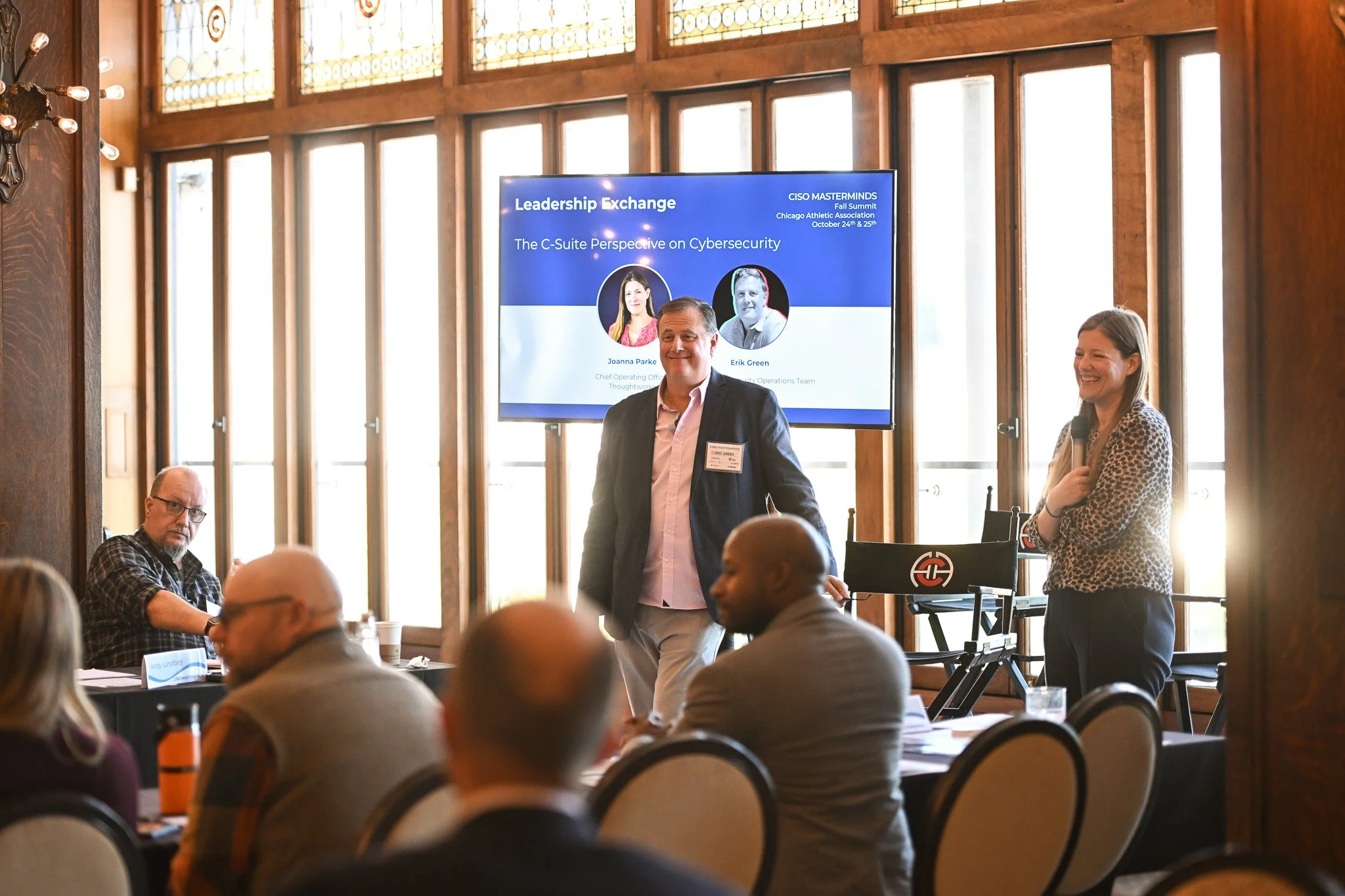 A professional conference room with wooden paneling and large windows. A man in a blazer and light-colored pants is speaking at a panel, with a woman beside him holding a microphone. A group of attendees is seated at round tables, listening to the pr