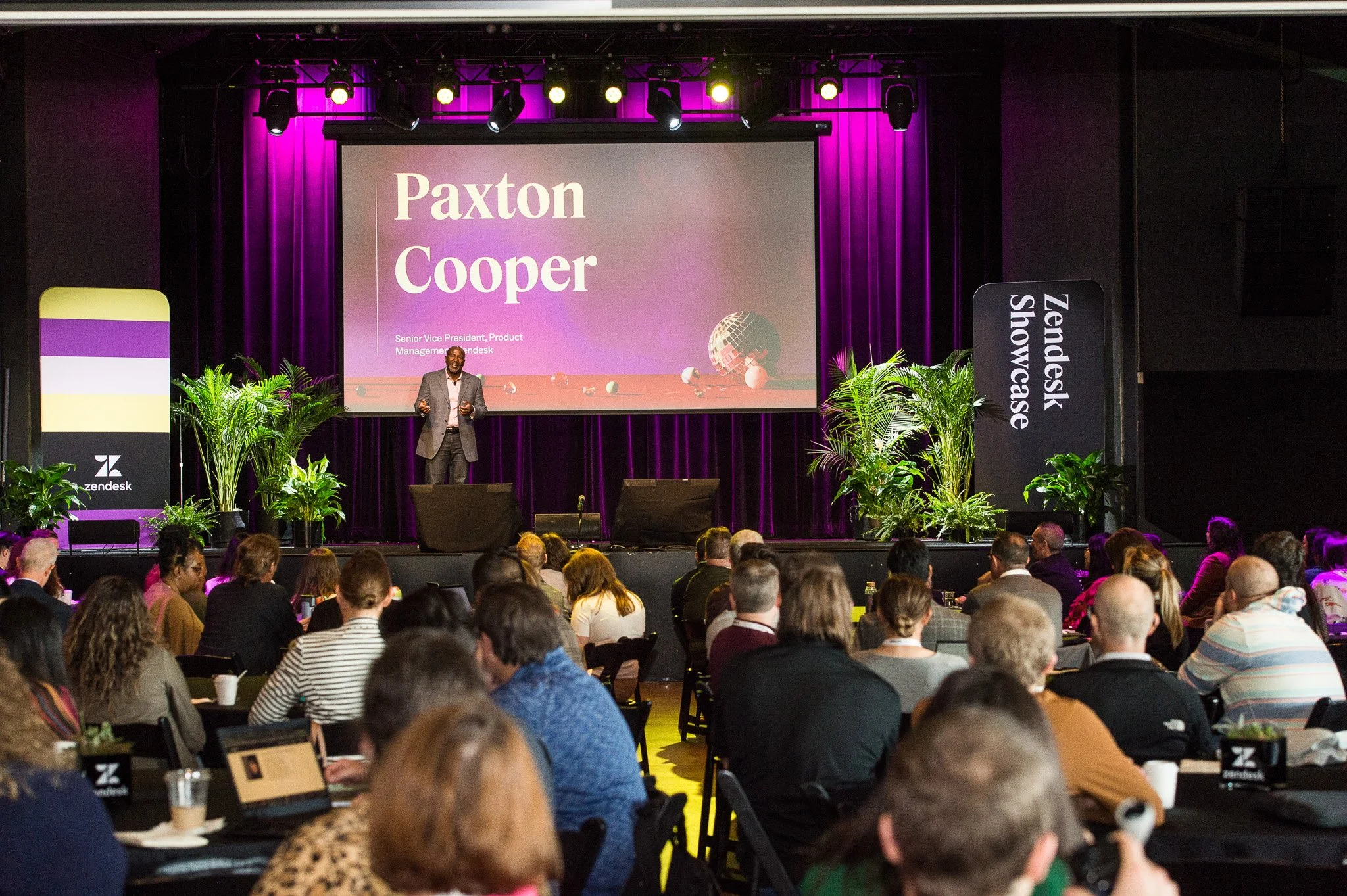 A speaker on stage at Zendesk Showcase event presenting to an audience. The stage includes a large screen displaying the name Paxton Cooper and his title as Senior Vice President, Product Management at Zendesk. The backdrop is purple, with banners on