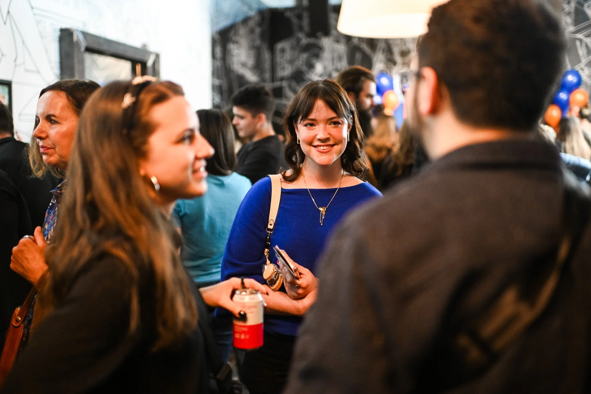 Group of people at an indoor social event, including a woman in a blue top smiling at a man with glasses and a beard, surrounded by other attendees, some with balloons in the background.