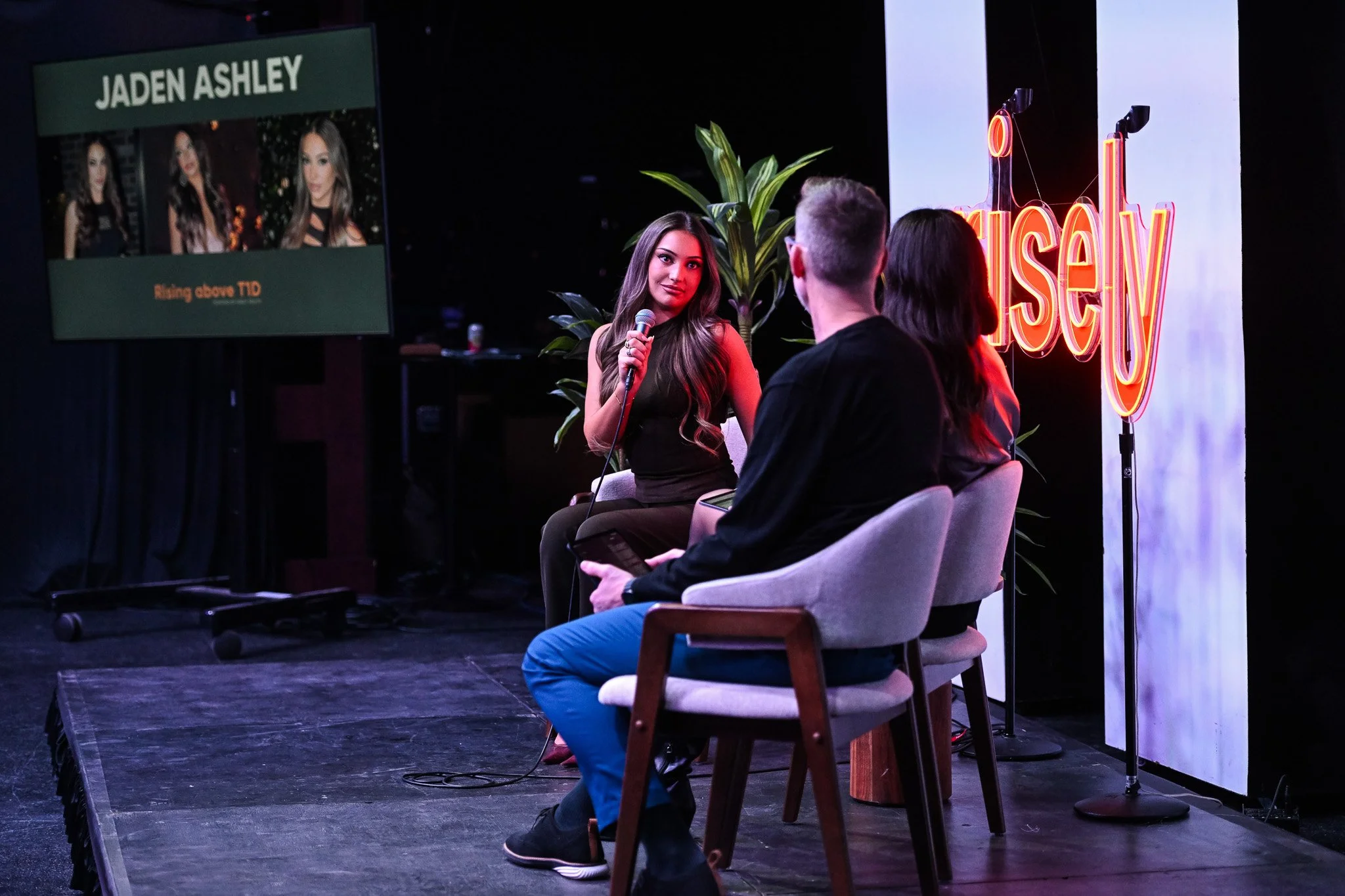 A woman with long brown hair sitting on a stage, holding a microphone, in conversation with two other people sitting in chairs; a large screen shows her picture and name, and a neon sign with part of the word 'Ours' is behind her.