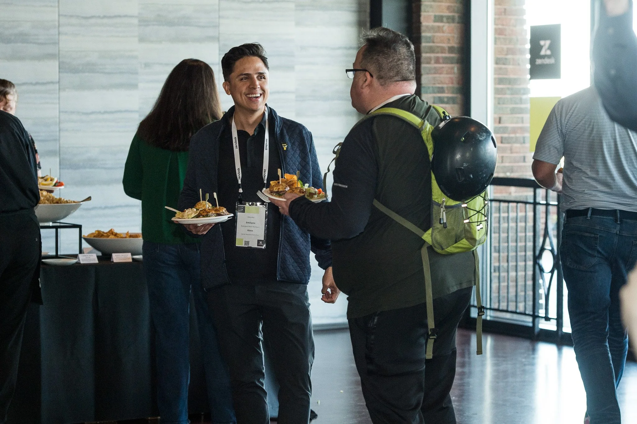Two men talking and smiling at a conference, holding plates of food, with food buffet table in the background.