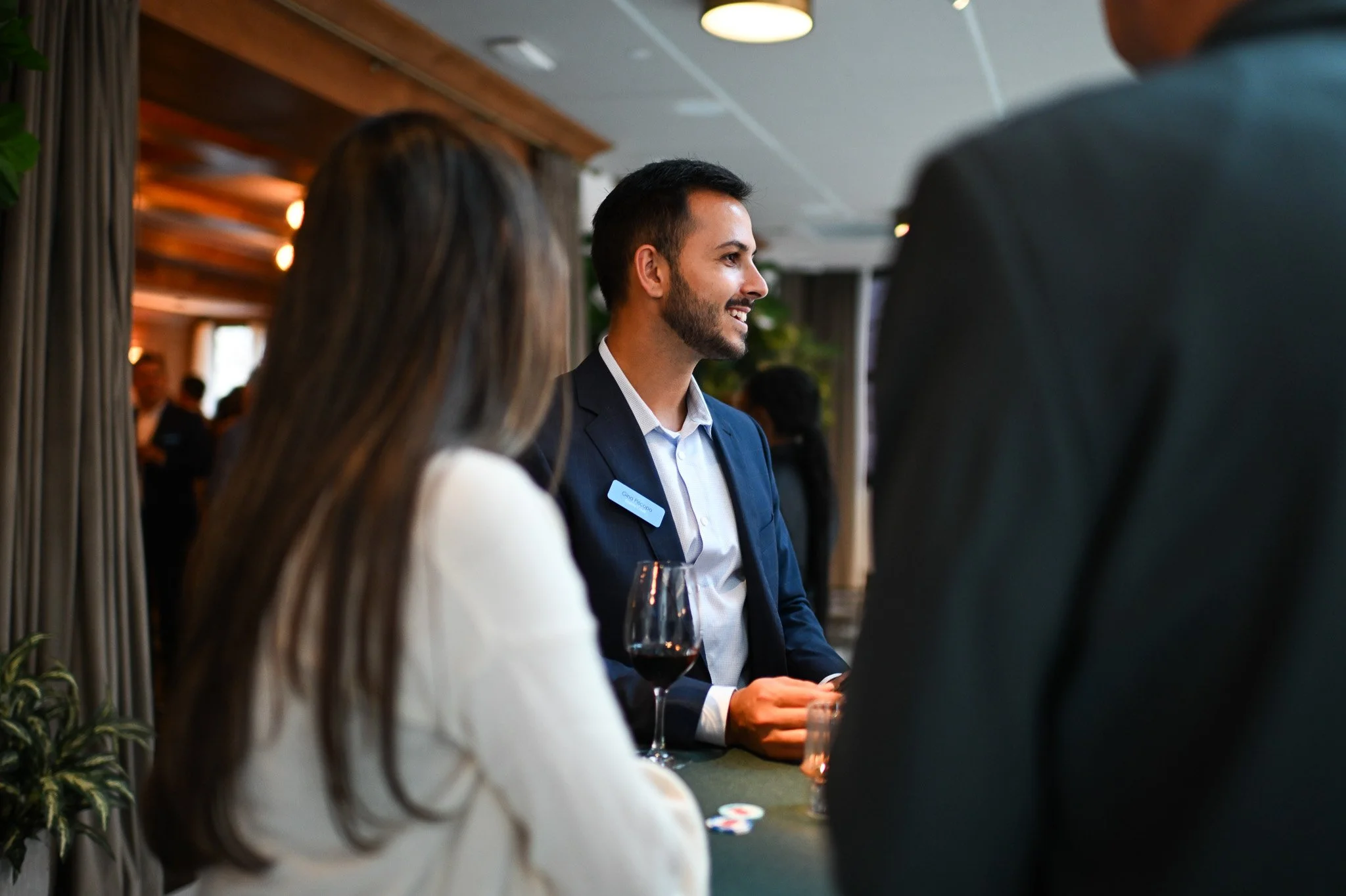 A bartender with a beard, dressed in a suit jacket and light shirt, smiling and talking to guests at a bar in a social setting.