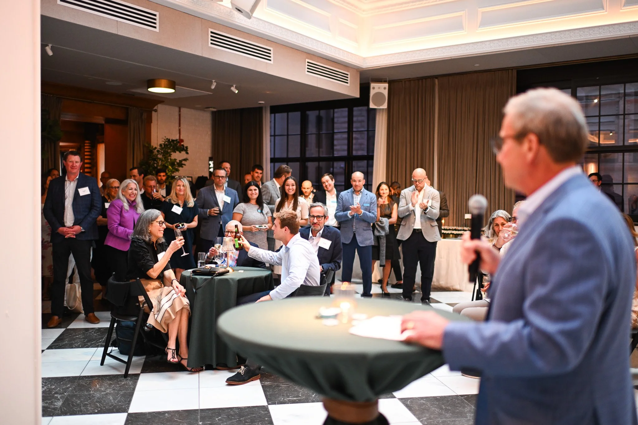 A man in a blue suit giving a speech at a business or social event, with a group of diverse people listening, smiling, and clapping in a well-lit room with large windows and checkered black-and-white floor.