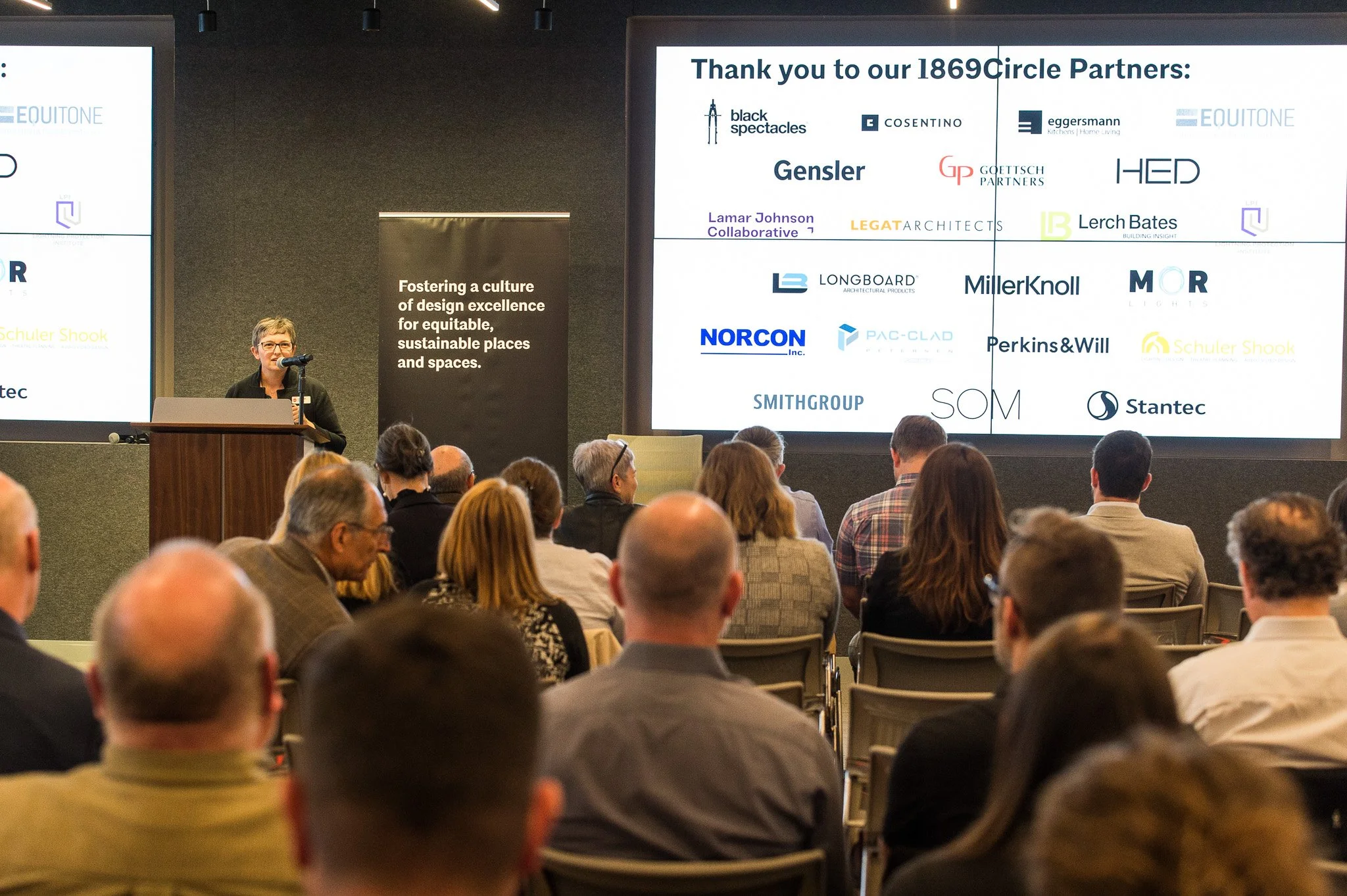 A woman giving a presentation at a conference, standing behind a podium with a microphone. A large screen behind her displays logos of various companies under the header "Thank you to our 1869 Circle Partners." An audience seated in front listens att