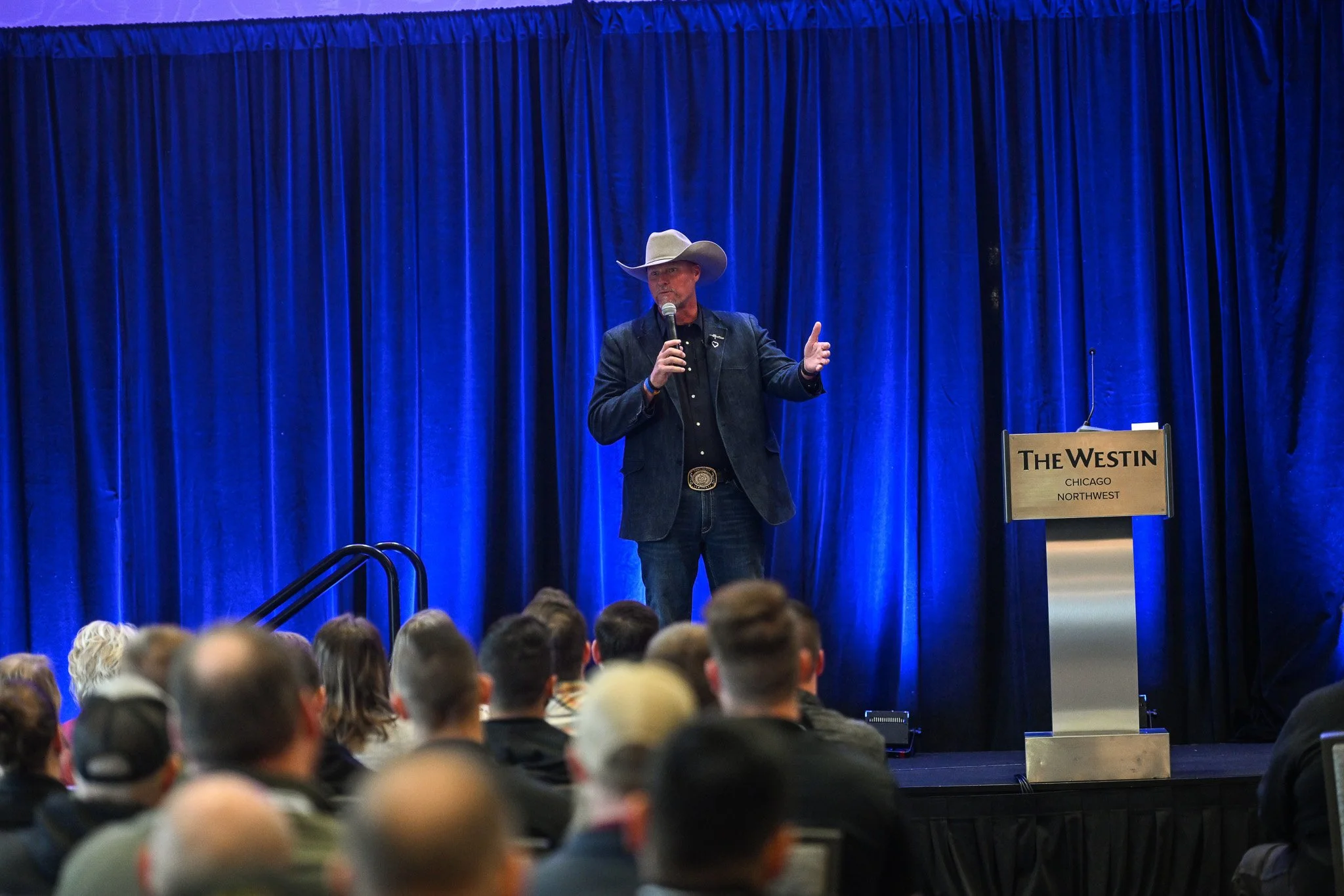 A man dressed in a suit and cowboy hat speaking into a microphone on stage at The Westin Chicago Northwest conference center, with an audience seated in front of him.