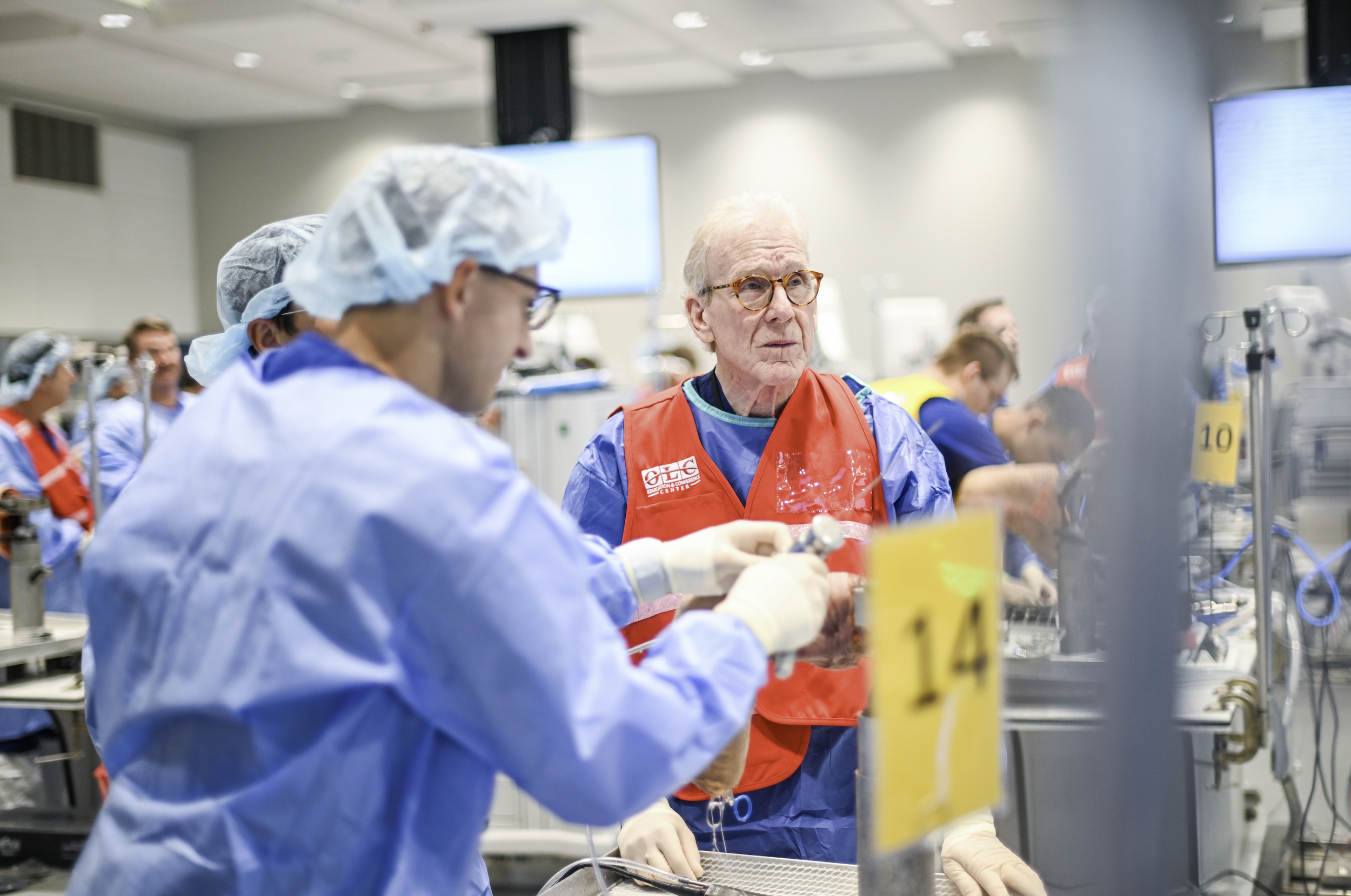 Medical professionals wearing scrubs, gloves, and hair covers working in a hospital or medical training environment. An elderly man with glasses in a red vest appears to be observing or instructing.