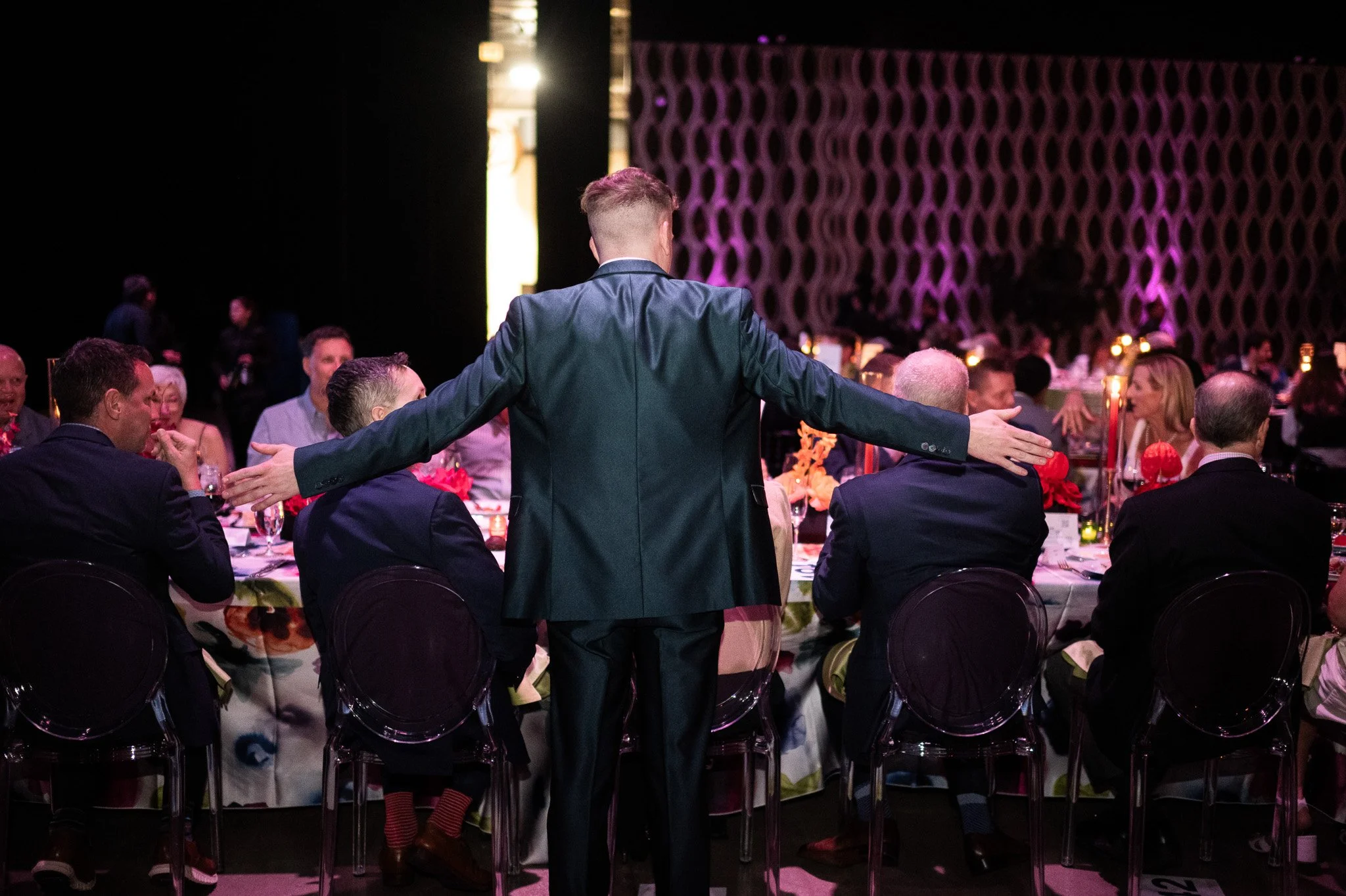 A man in a suit with arms outstretched standing behind a seated man at a banquet table during a formal event with many other guests, food, and decorations.