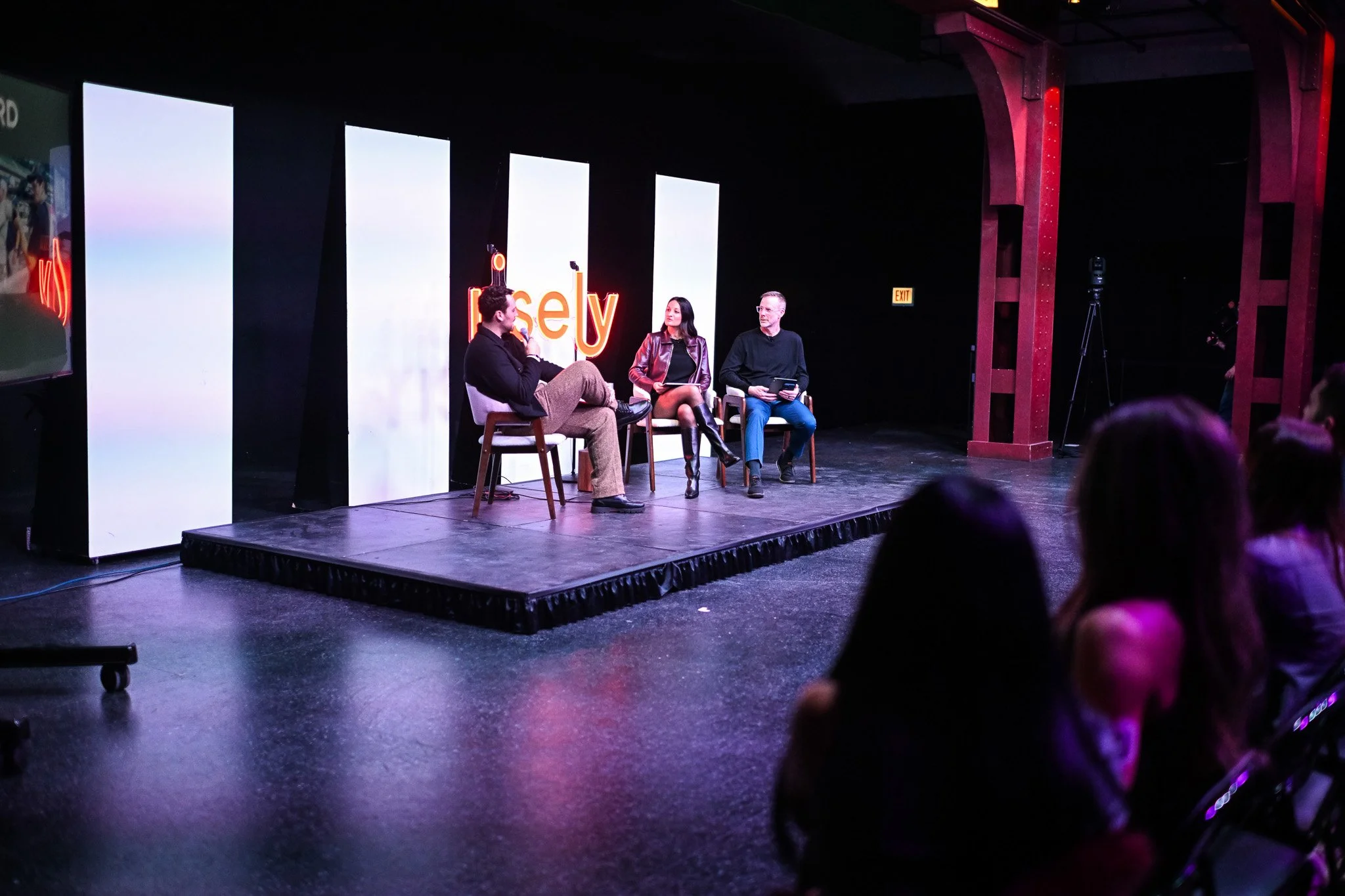Three people sitting on chairs on a small stage, participating in a panel discussion with a screen behind them displaying the word 'Trust' in orange letters, and an audience watching in front of them.