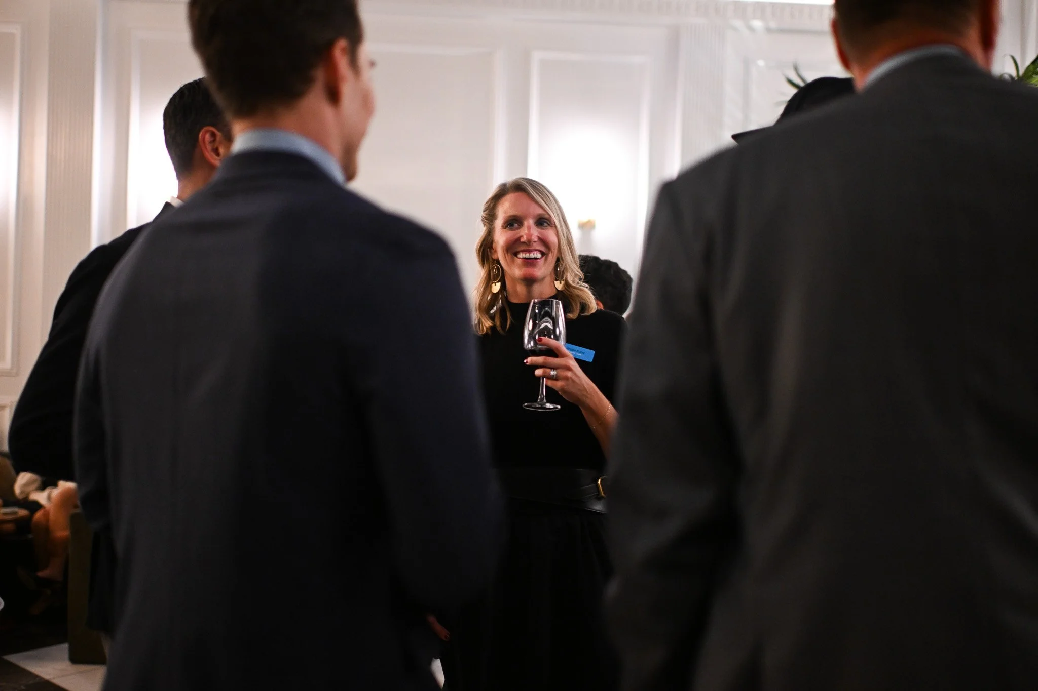 A woman smiling and holding a glass of red wine, engaged in conversation with a group of men in business suits at a social event indoors.