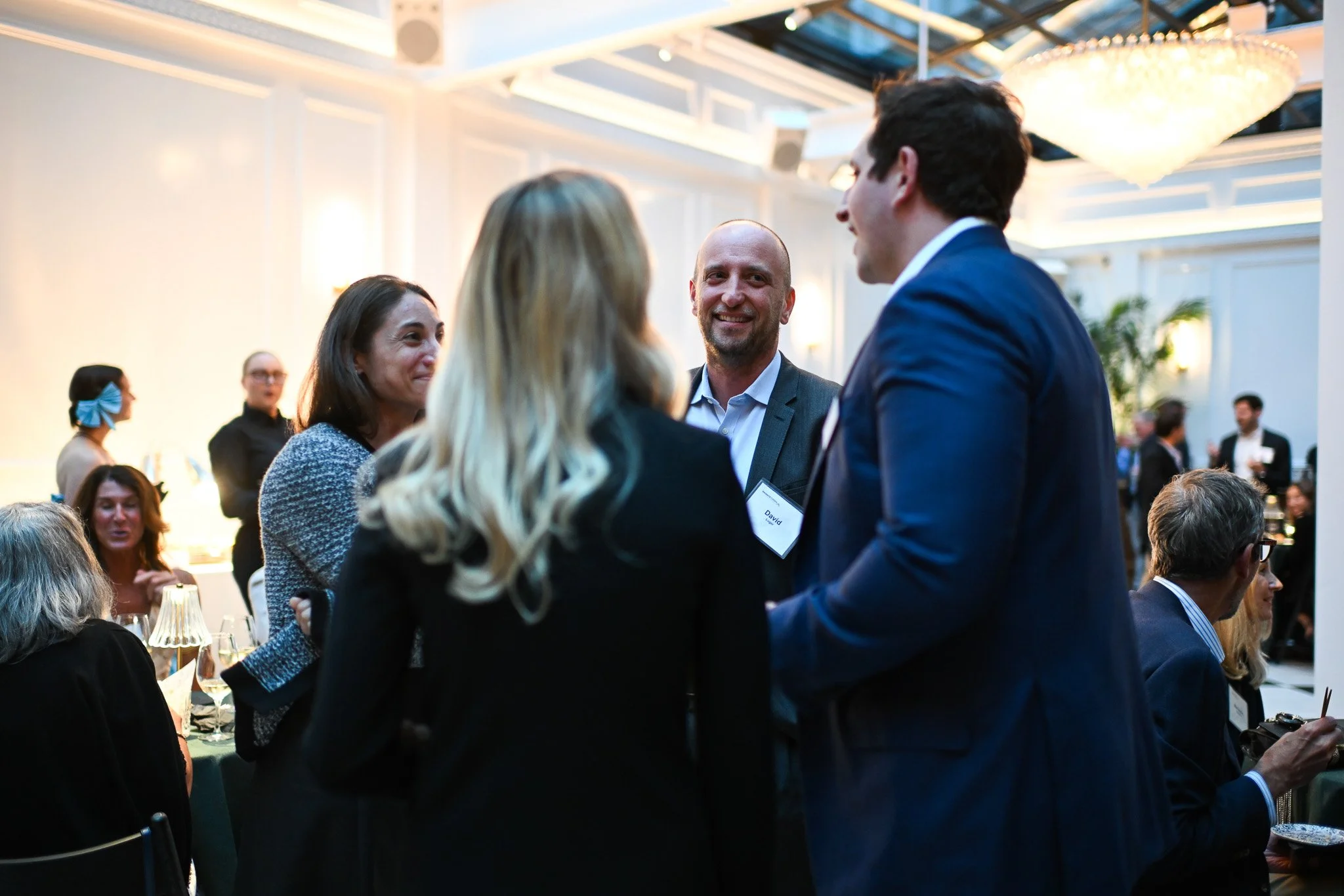 A group of well-dressed people at a formal event engaging in conversation, with a man in a gray suit smiling at a woman in a black blazer.