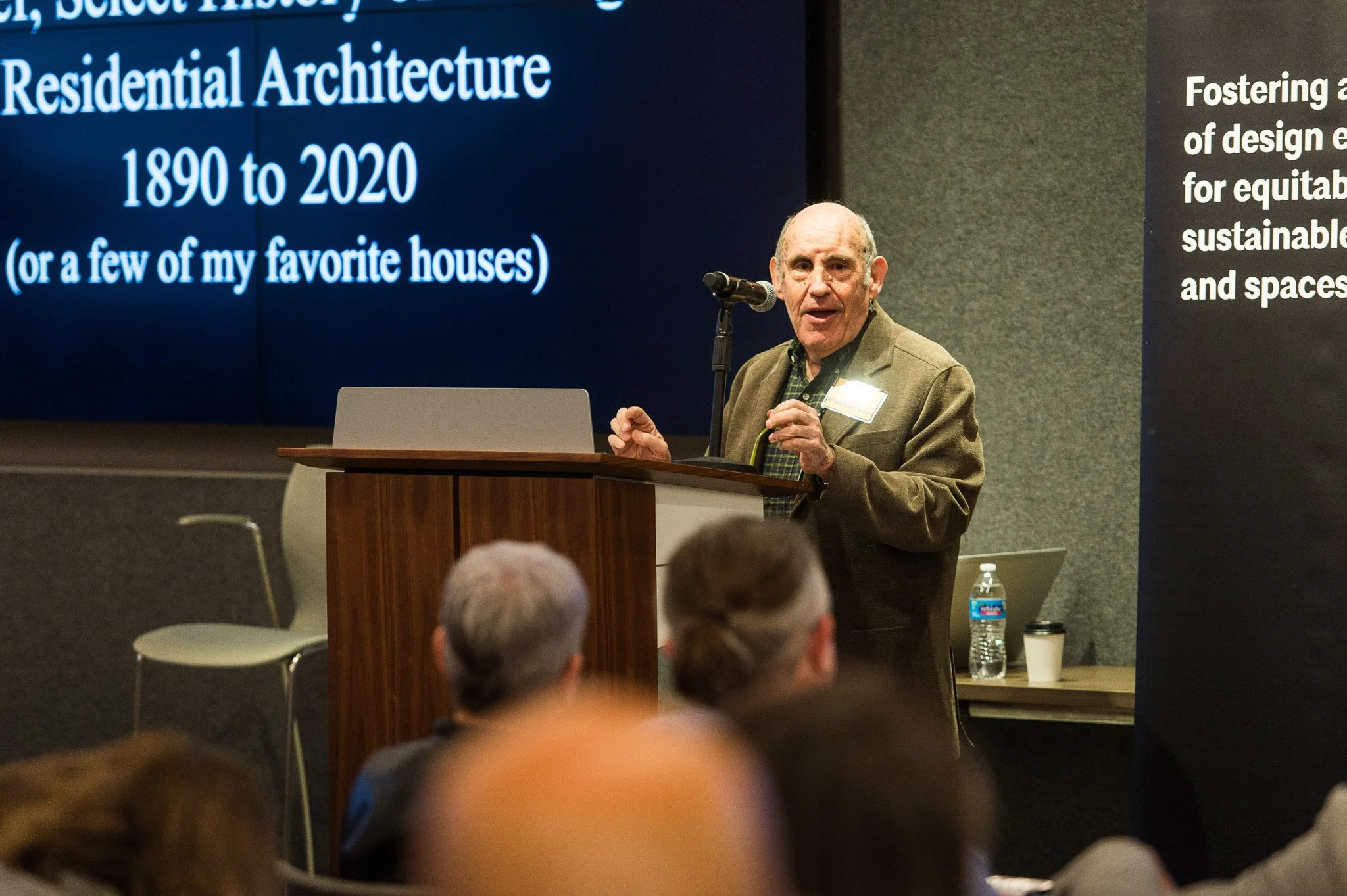 An elderly man giving a speech at a conference, standing behind a podium with a microphone. A large screen behind him displays a presentation titled 'Residential Architecture 1890 to 2020' with a subtitle '(or a few of my favorite houses)'. Several a
