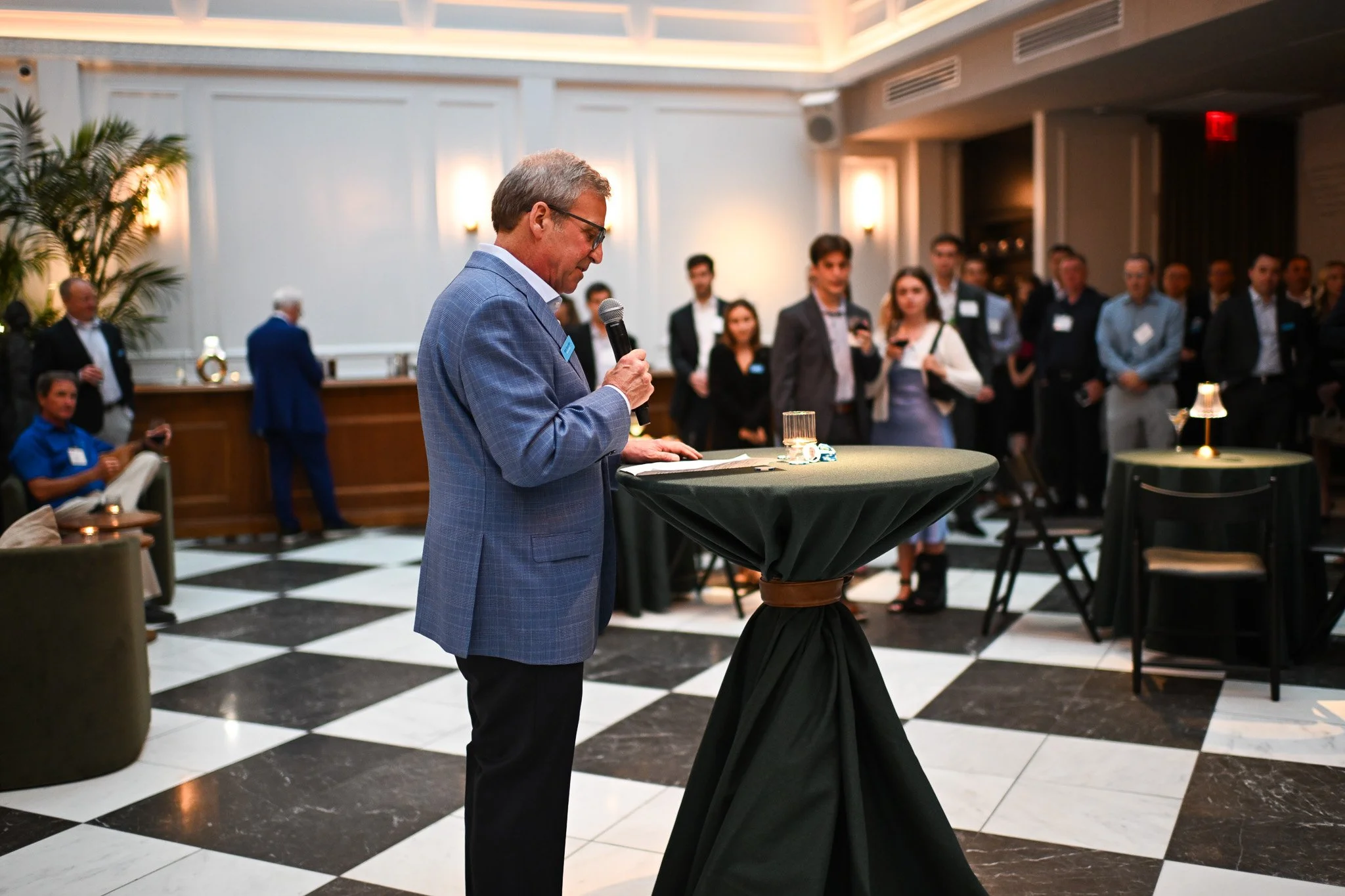 A man in a blue checkered suit holding a microphone and reading from a paper at a round, high-top table in a formal event space. Several people are standing and sitting in the background, listening and engaging with each other.