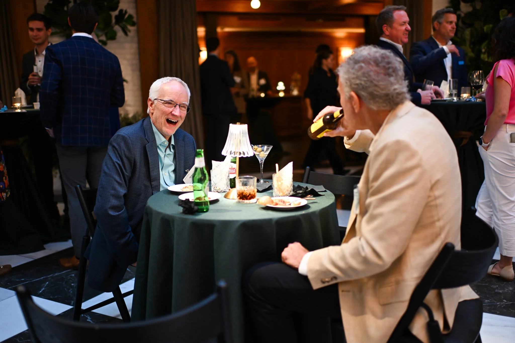 People socializing at a lively gathering or party, some sitting at a table, others standing and chatting, in an elegant indoor setting.