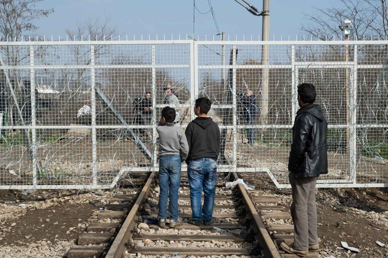 Idomeni, Greece – March 1, 2016 Refugees look through a border gate into Macedonia (FYROM). An active railway, largely for freight trains, runs between Greece and Macedonia (FYROM). Armed guards patrol the crossing on the Macedonian side.
