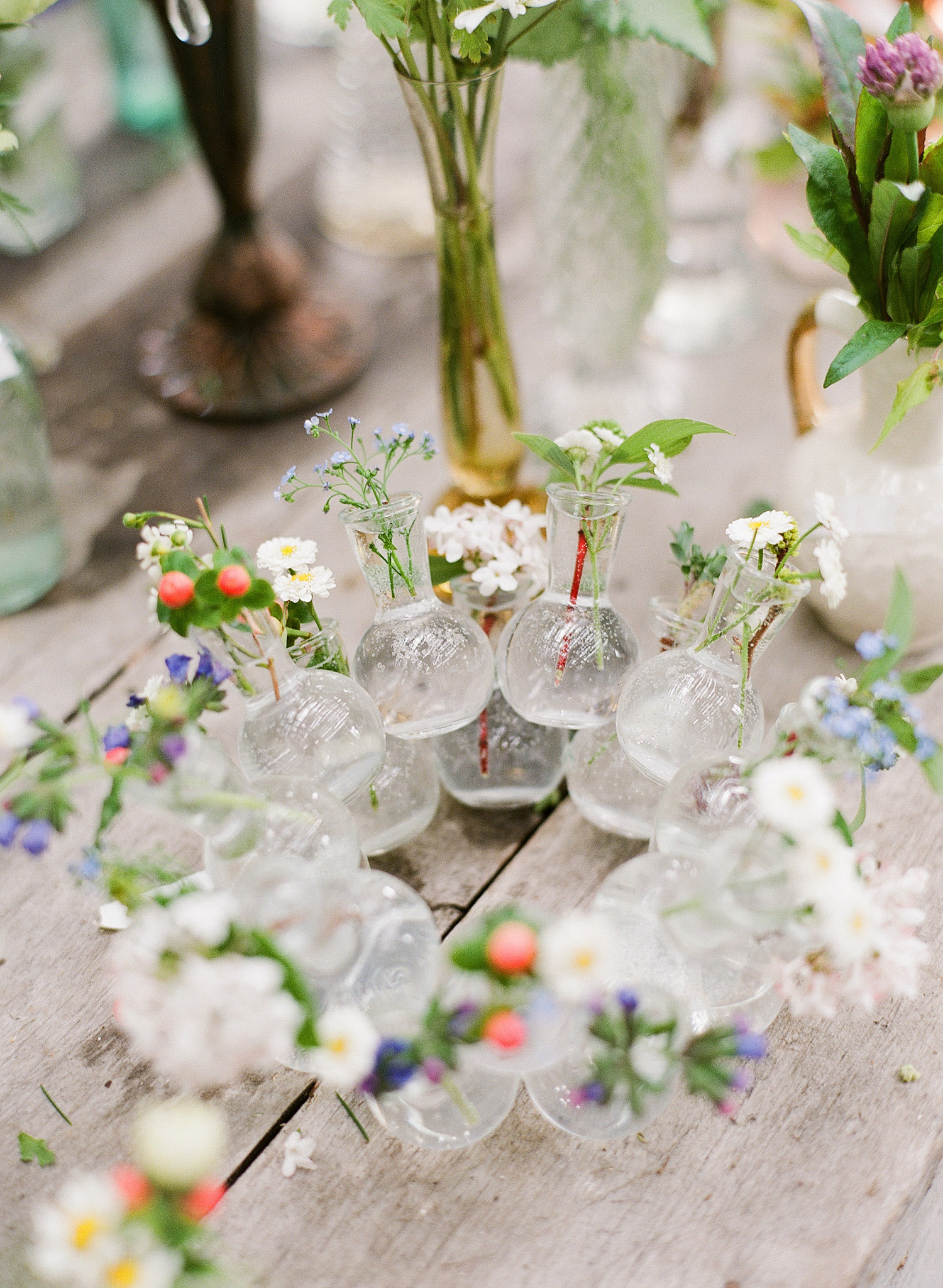 a wreath of vases with springtime buds sits on a farm table while destination wedding photographer Lexia Frank photographs this springtime wedding on film at the Northwind Perennial Gardens in wisconsin