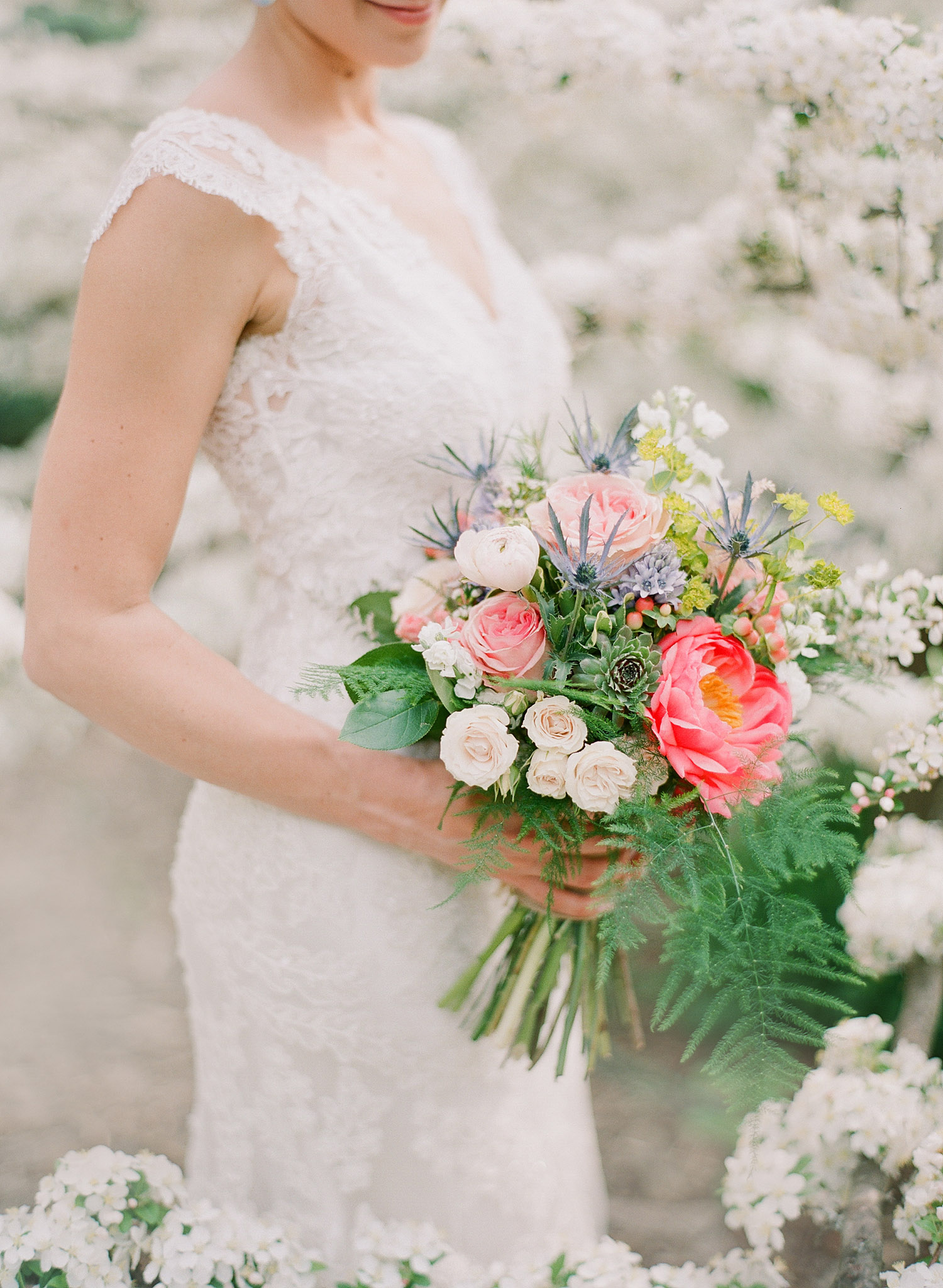 Bride holds asymetrical bouquet of terms and ranunculus succulents and roses as she stands in the springtime blooms of Northwind Perennial Gardens in wisconsin while Lexia Frank, destination wedding photographer, photographs on film 