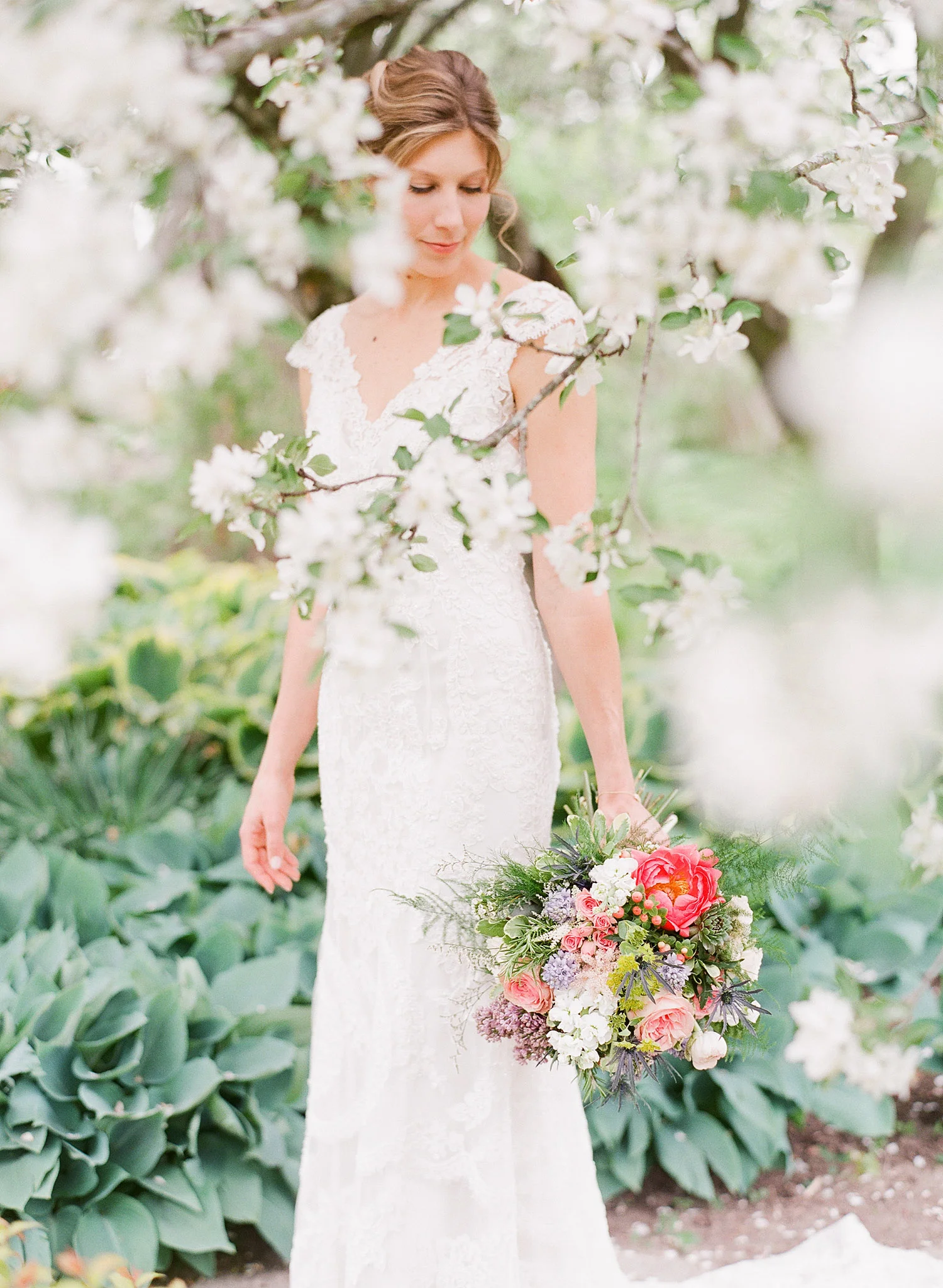 the bride stands among blooming trees and hostas holding her asymmetrical bouquet of ranunculus peonies terms lavender and roses at her springtime wedding at Northwind Perennial Gardens - a beautiful wisconsin wedding venue- while Destination Wedding Photographer Lexia Frank photographs her wisconsin wedding on film 