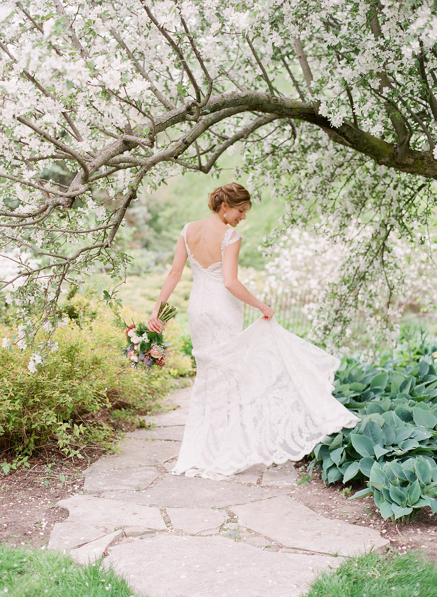 bride twirls her lace wedding dress with cap sleeves under blooming trees at her springtime wedding at Northwind Perennial Gardens - a beautiful wisconsin wedding venue - while destination wedding photographer Lexia Frank photographs her wedding on film 
