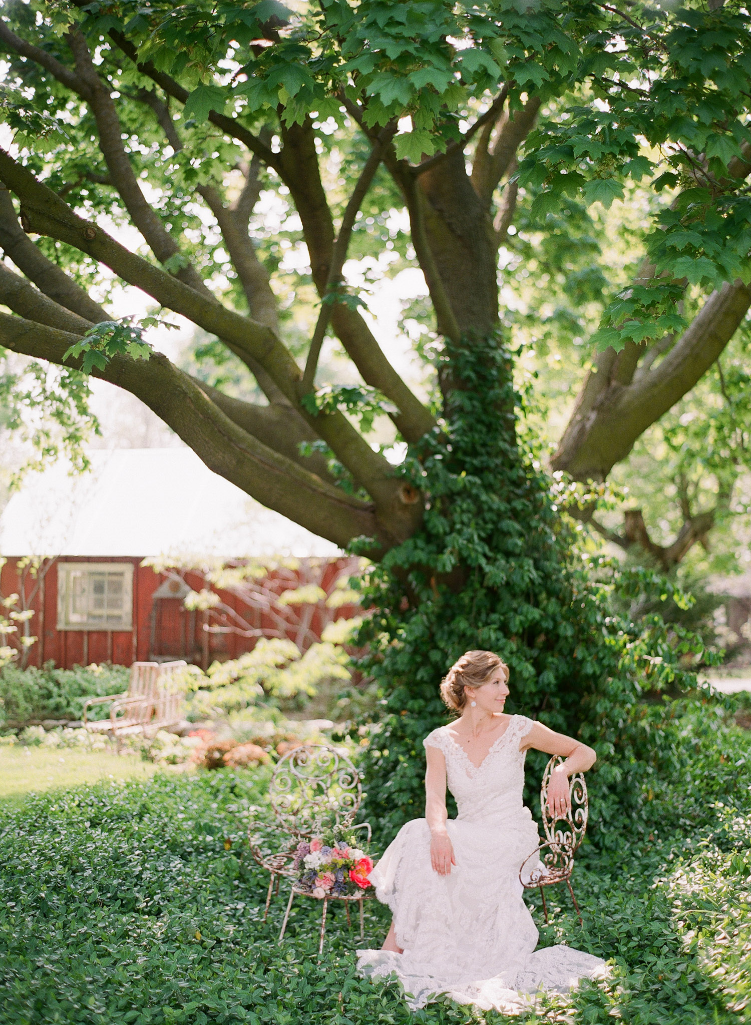 bride sits on a vintage wrought iron chair in ivy under a tree at this springtime wedding at Northwind Perennial Garden - a beautiful wisconsin wedding venue - while Destination wedding photographer Lexia Frank Photographs this wedding on film