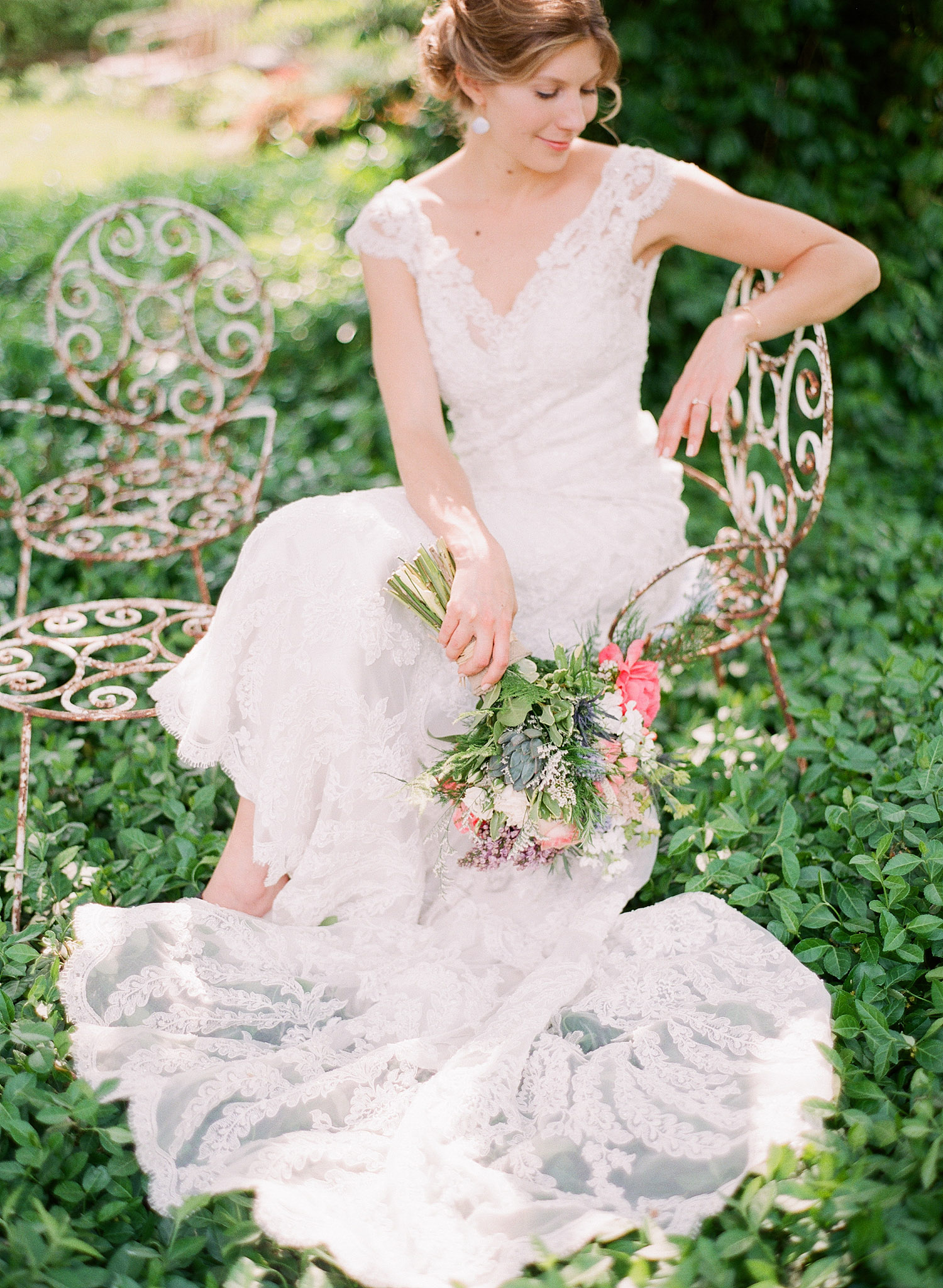 bride sits on vintage wrought iron chair in ivy in her vintage lace wedding dress with cap sleeves holding her asymmetrical spring wedding bouquet at this springtime wedding at Northwind Perennial Garden - a beautiful wisconsin wedding venue - while Destination wedding photographer Lexia Frank Photographs this wedding on film