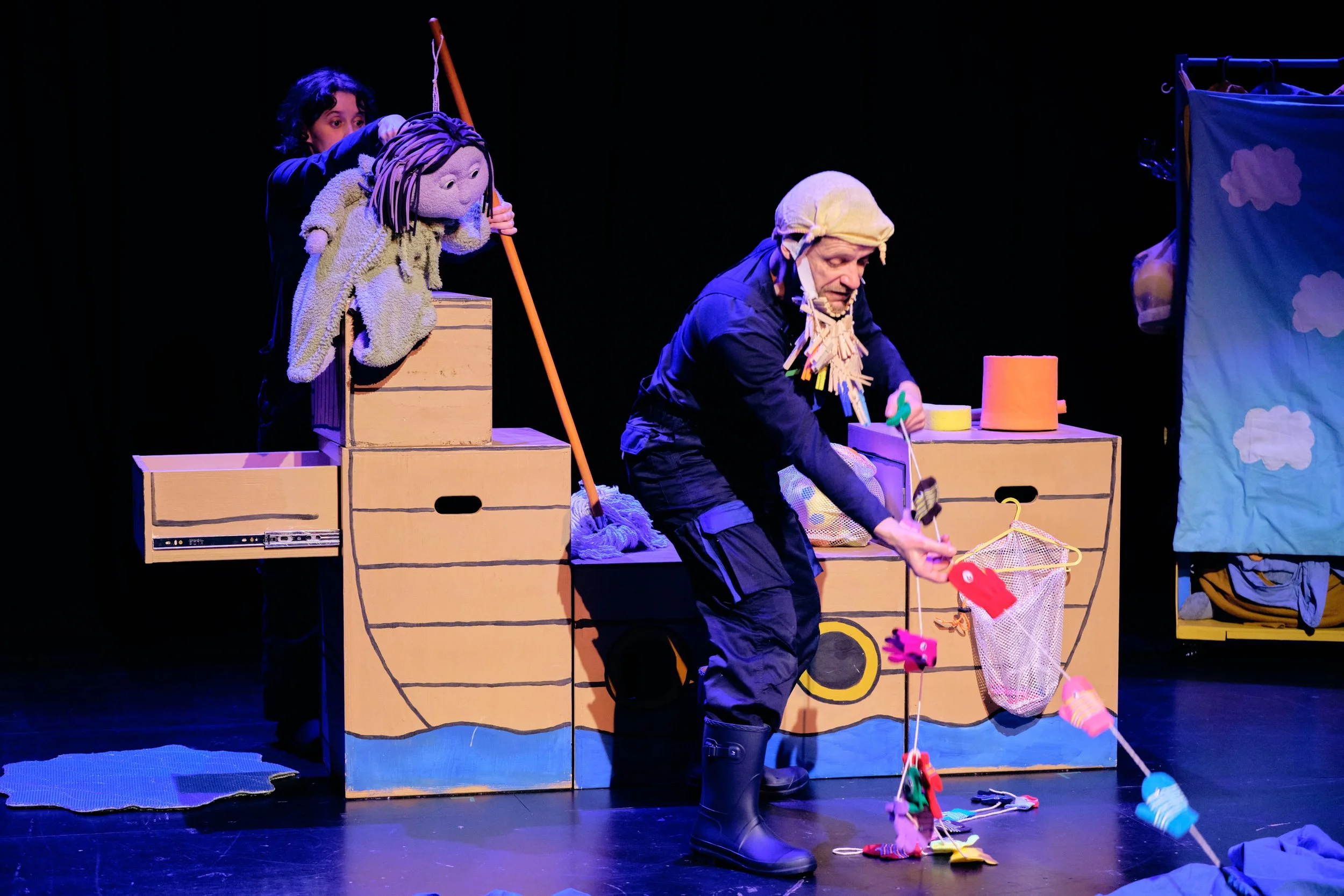 Puppeteer and costumed performer act in a colorful theatrical set with puppet, cardboard boxes, and fishing props.