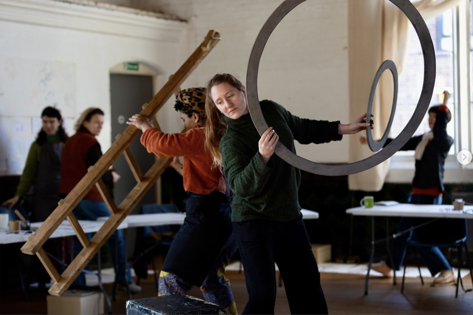 Artists in a workshop moving large wooden structures and metal rings during a collaborative performance exercise.