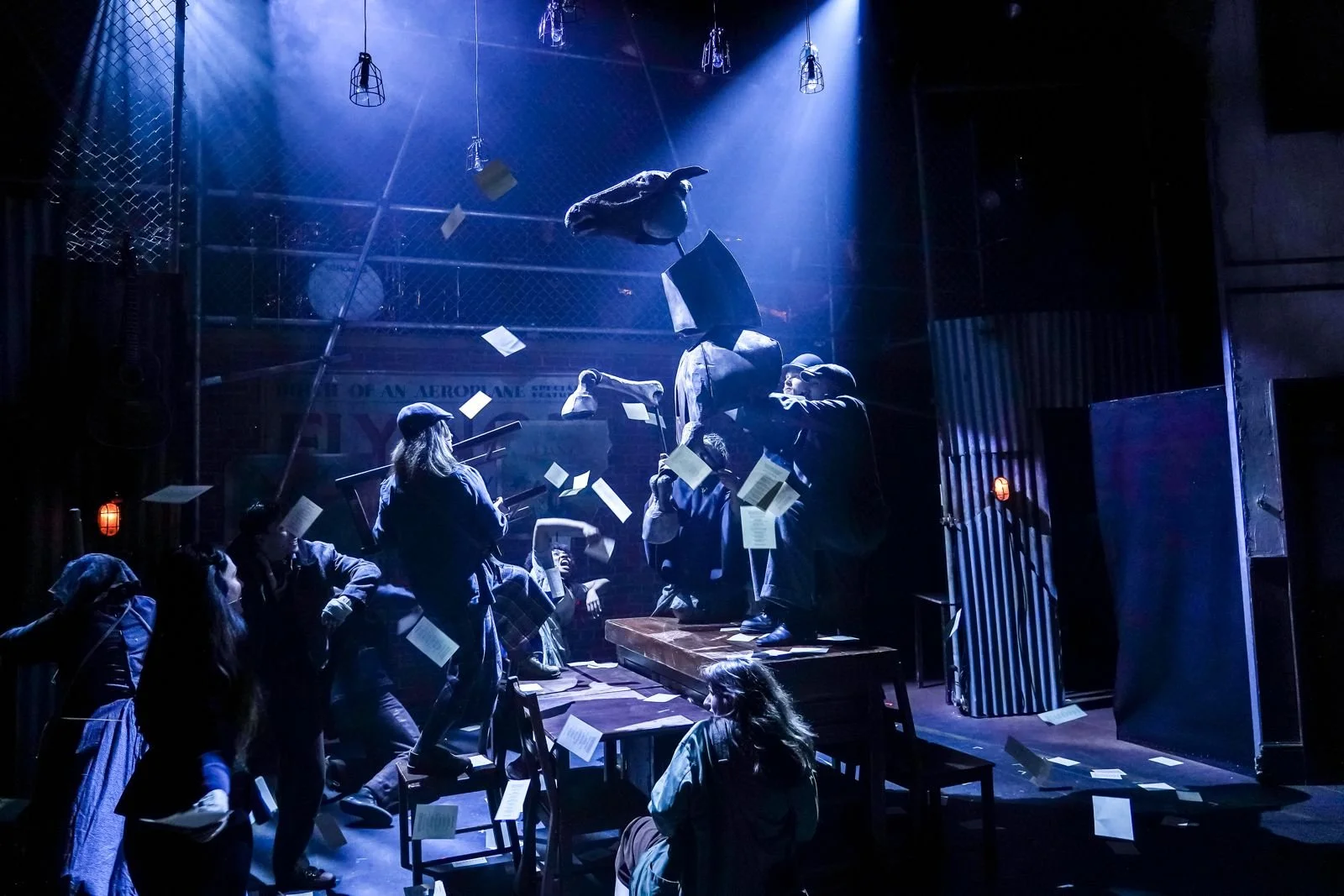 Dark theatrical stage scene with actors, floating papers, blue spotlights, cages, and a suspended horse puppet