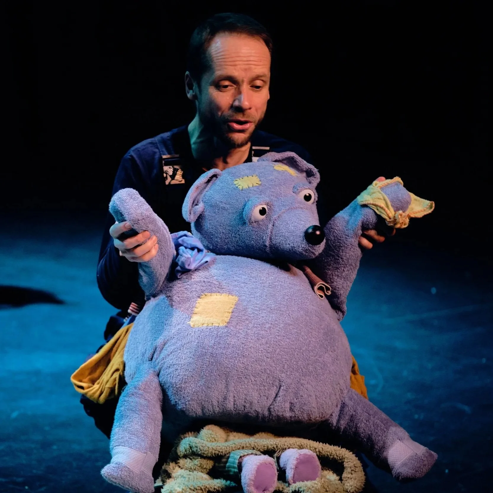 Puppeteer holding a large patched blue bear puppet on a dark stage
