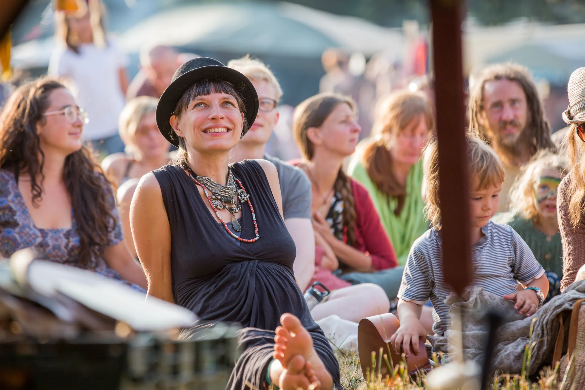 A woman with a black hat, short dark hair, and layered necklaces smiling and sitting barefoot at an outdoor event surrounded by other people, some with face paint and colorful clothing.