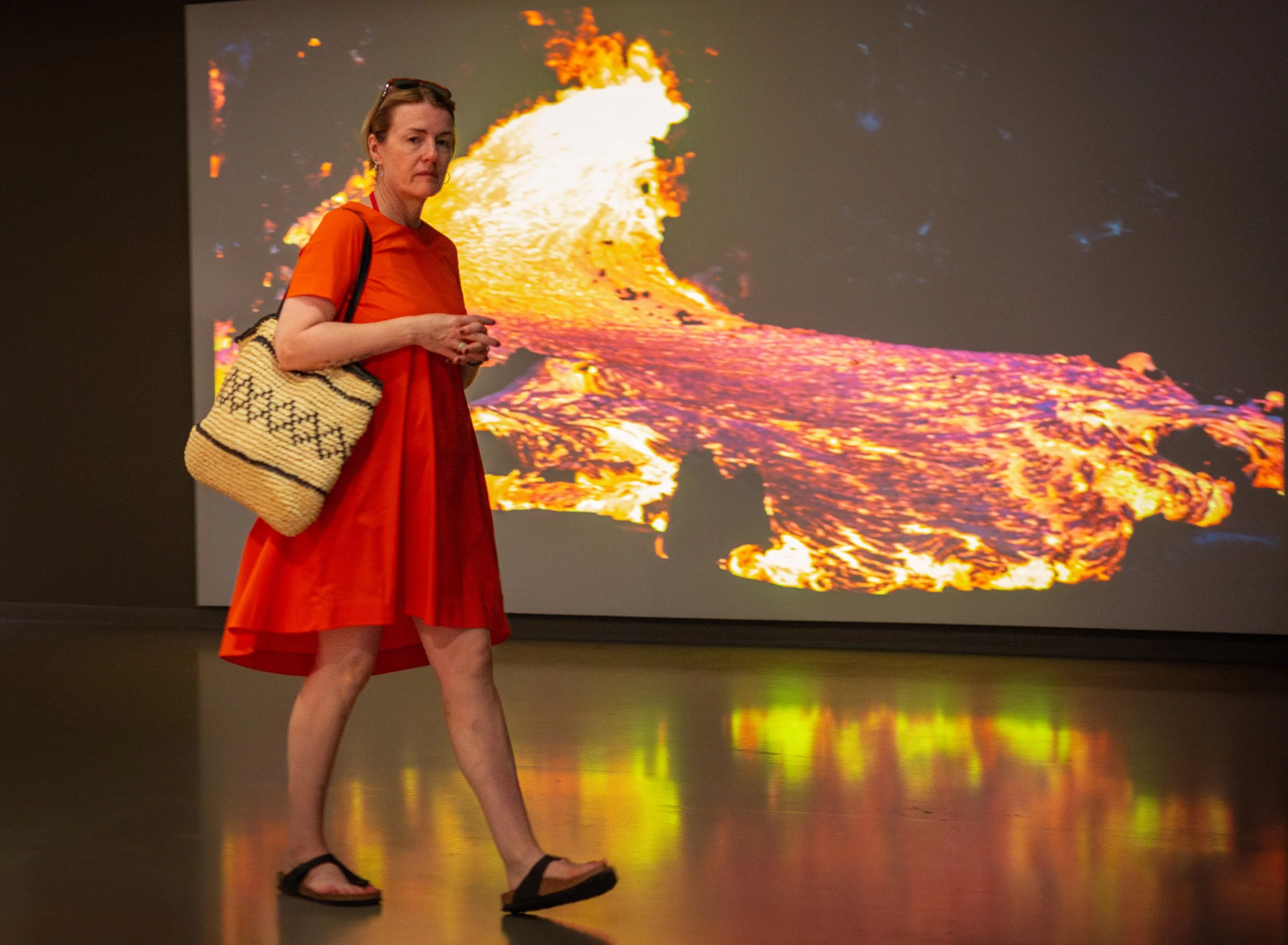 A woman in an orange dress with a black-and-white woven bag walks inside an art gallery, with a vibrant volcanic eruption image displayed on a screen behind her.