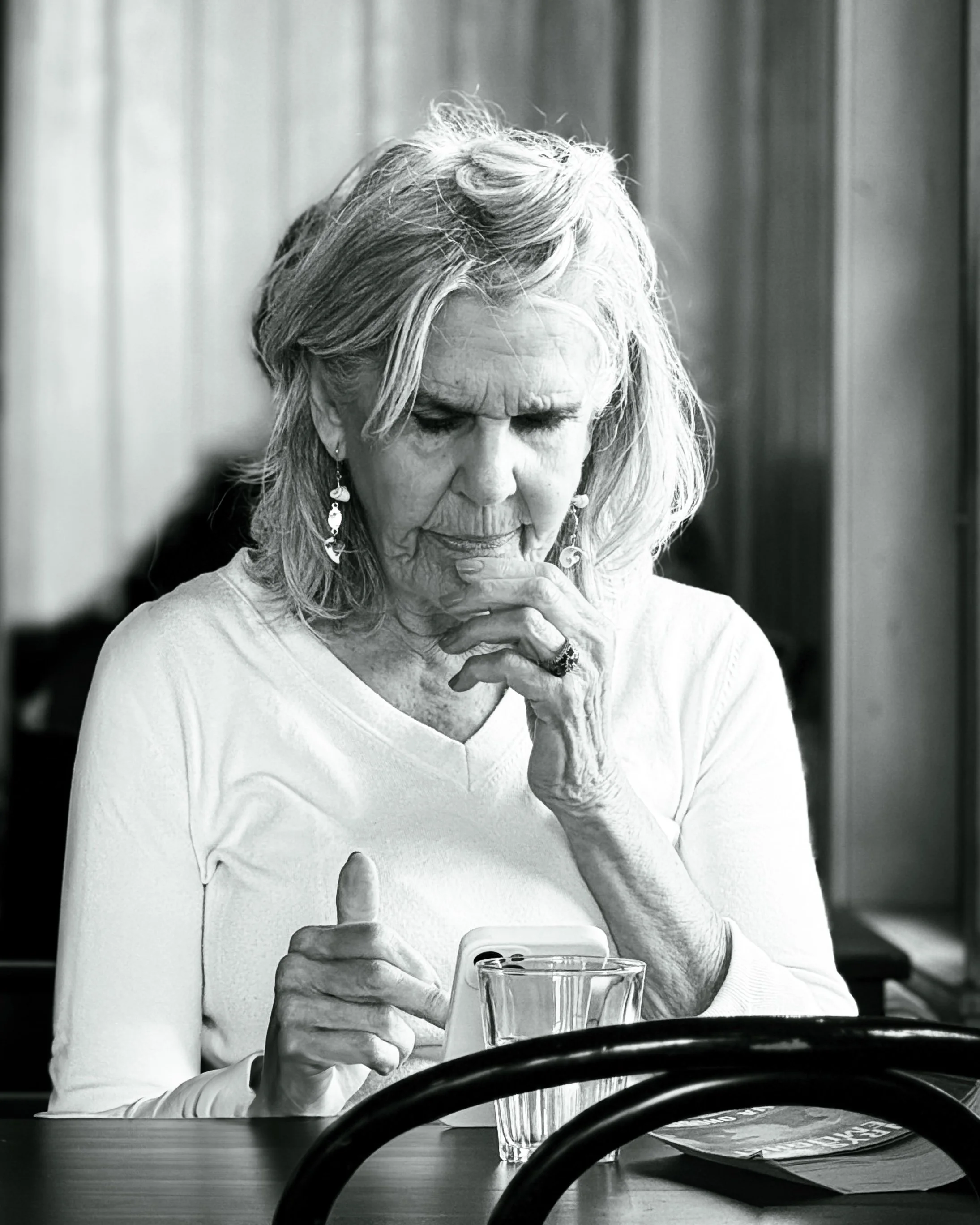 An woman with light hair wearing a white long-sleeve shirt, earrings, and a ring, sitting at a table, looking at a smartphone with a puzzled expression, with a glass of water and a magazine on the table in front of her.