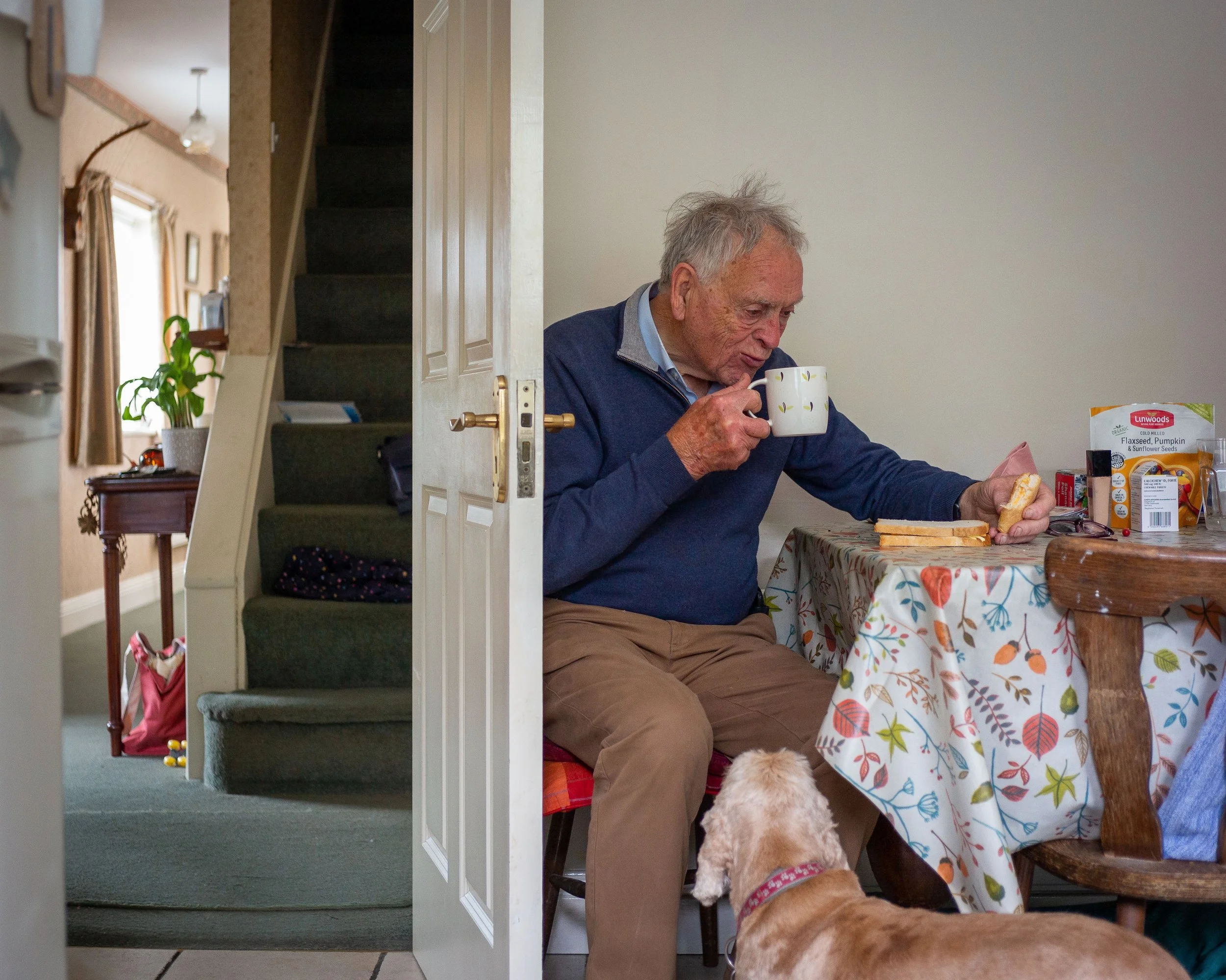 An elderly man sitting at a dining table, drinking from a mug with toast and food on the table, while a dog looks up at him. The room has a staircase and a window in the background.