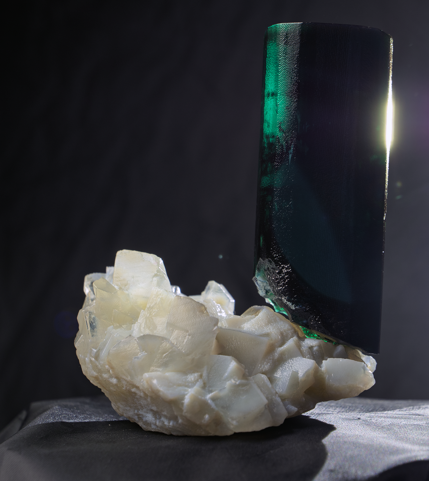 A rectangular, dark green and black mineral or gemstone with a shiny surface, partially embedded in a rough, white chalk-like mineral formation, against a dark background with light illuminating the edge of the gemstone.
