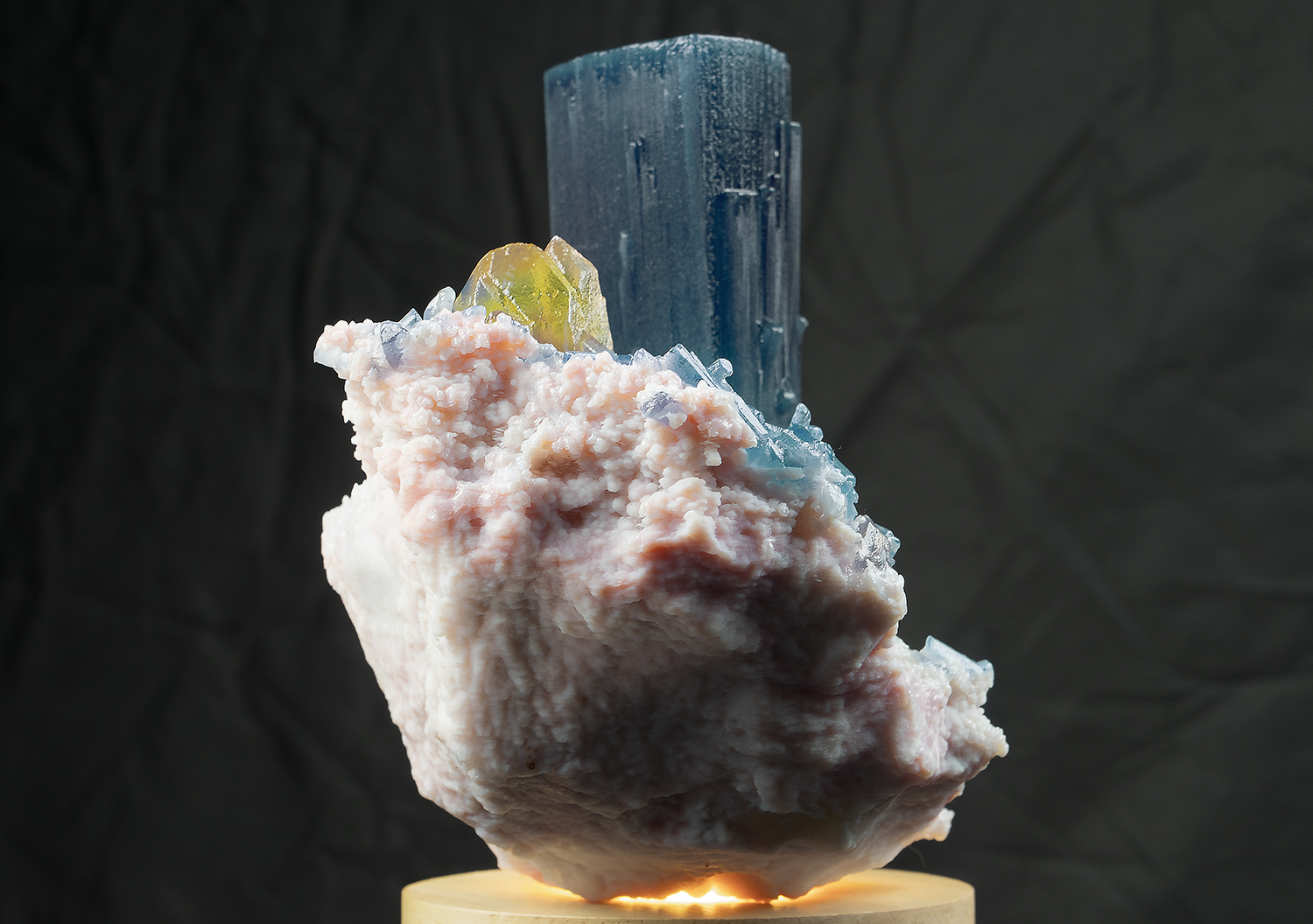 Close-up photograph of a mineral specimen with a large, rectangular blue crystal, yellowish-green mineral, and a pinkish-white, rough textured mineral base, illuminated from below and set against a dark background.
