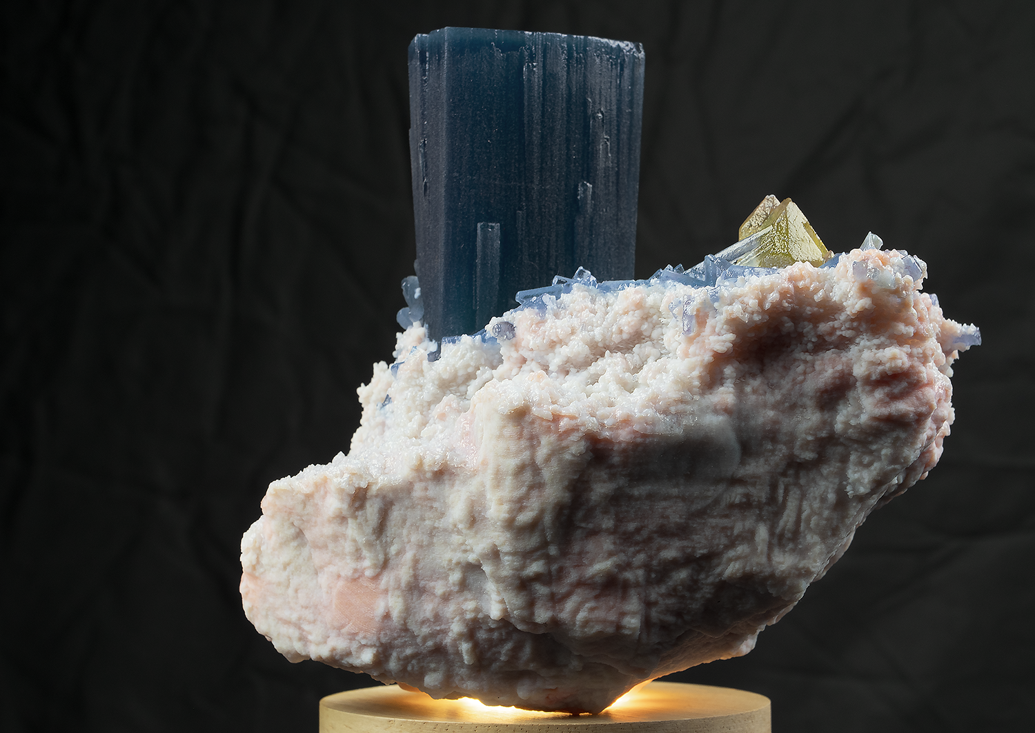 A large mineral specimen with a blue crystal pegmatite, light-colored boulders, and smaller crystals on a wooden stand against a dark background.