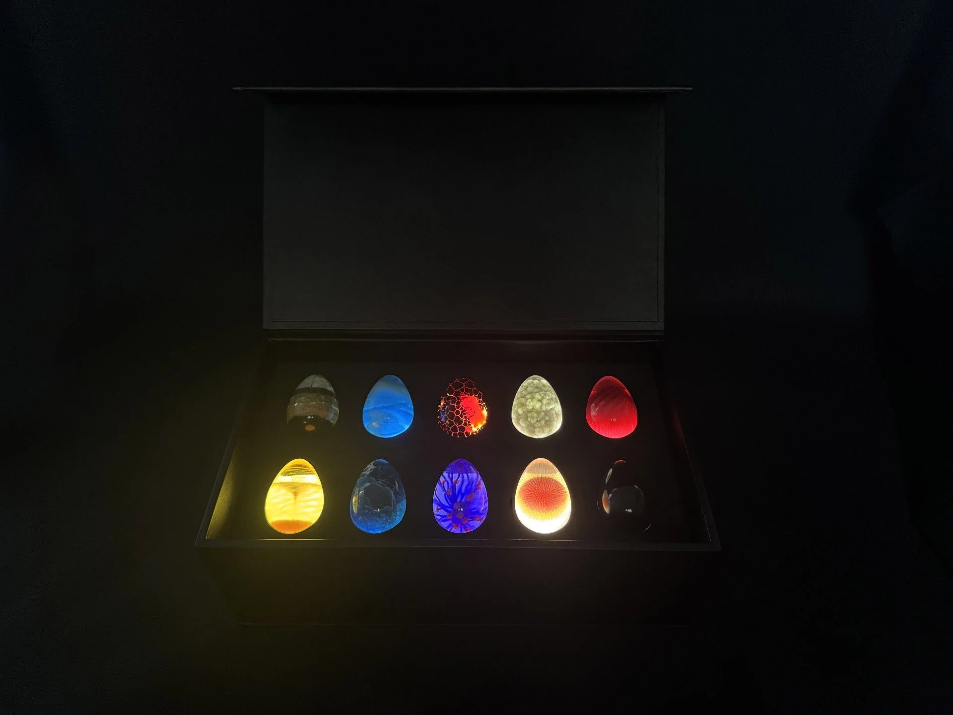 A black box with a glass top contains nine colorful glass eggs, each with unique patterns and colors, illuminated from within in a dark setting.
