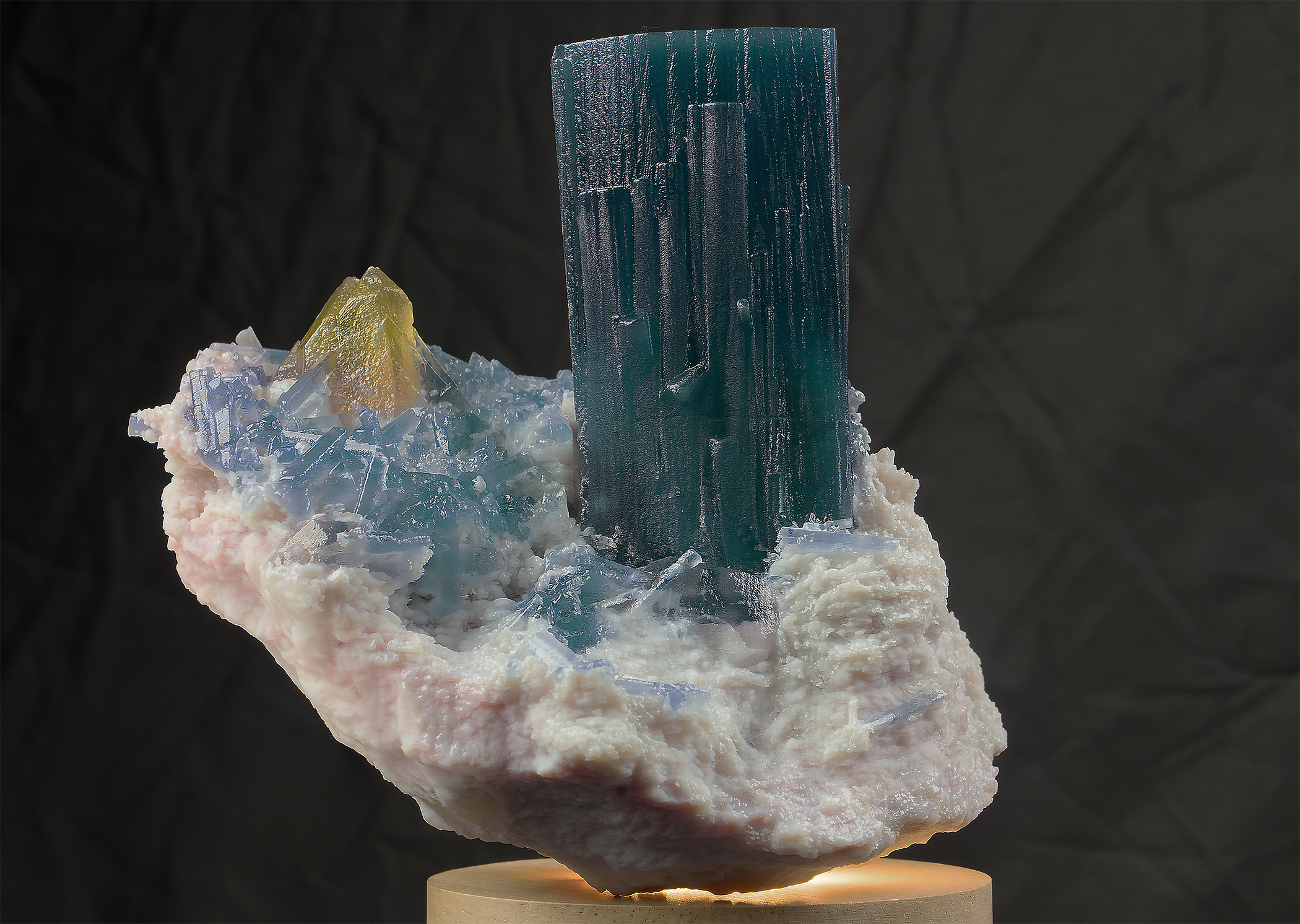 A mineral specimen featuring a large vertical blue-green crystal amid smaller clear and yellow crystals on a light-colored base, displayed on a round wooden stand with a dark background.