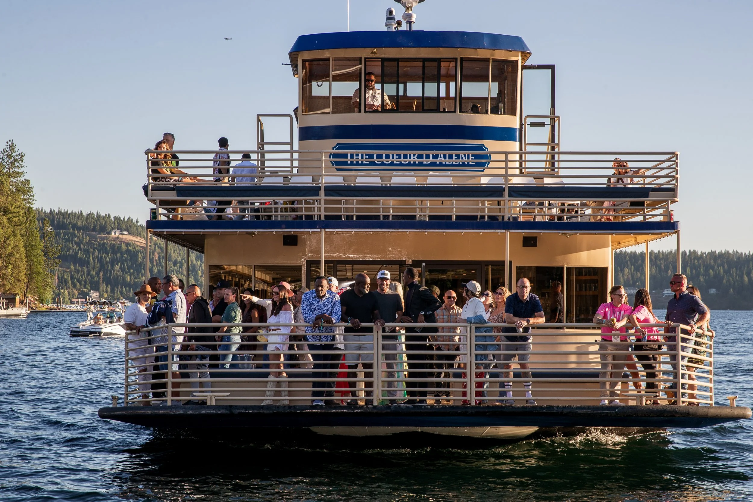 A large boat with multiple decks carrying many passengers, some on the lower and upper decks, on a body of water with trees and boats in the background during daylight.