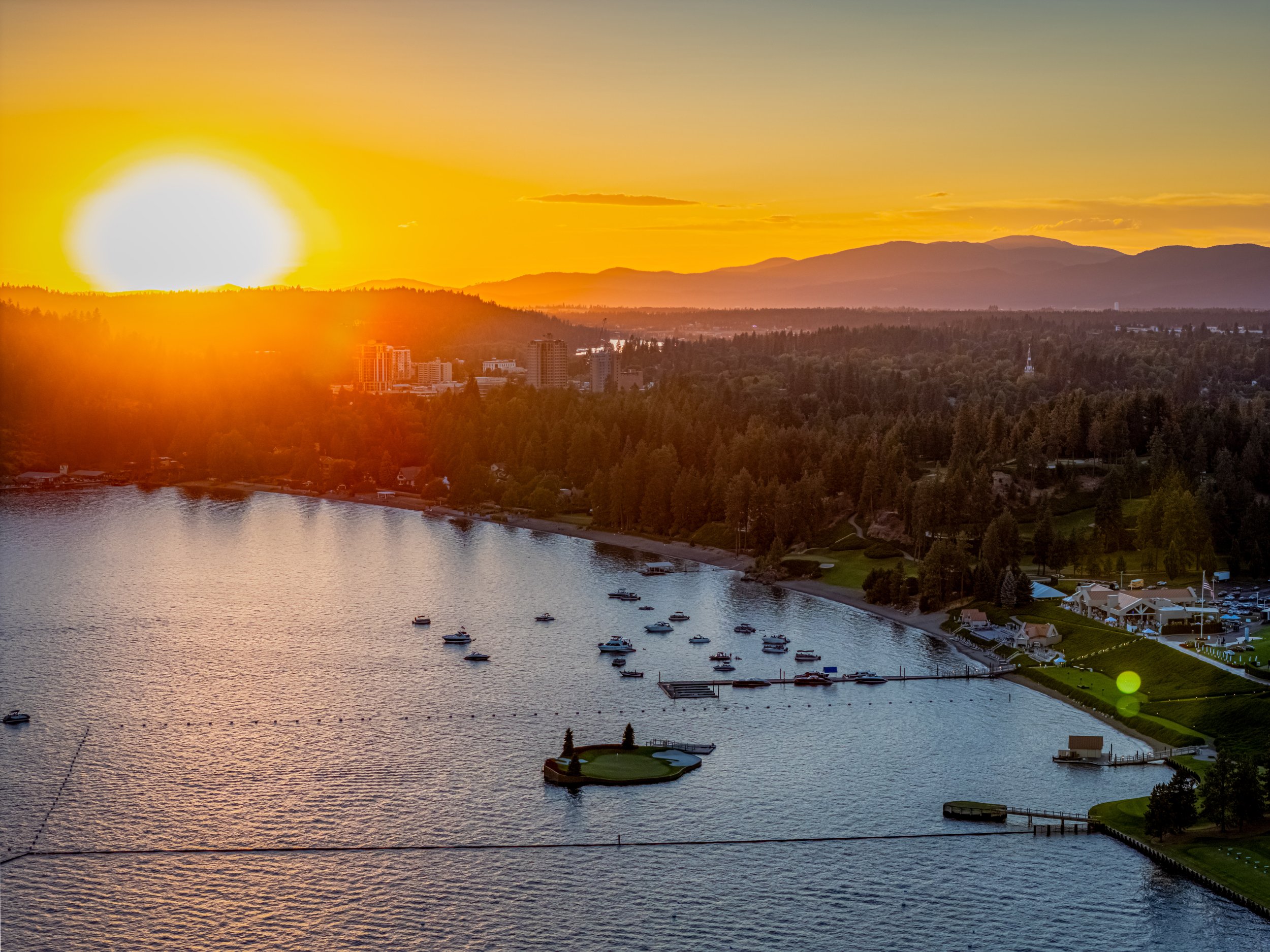 Aerial view of a waterfront city during sunset with boats on the water, surrounded by green trees and buildings, with mountains in the background.