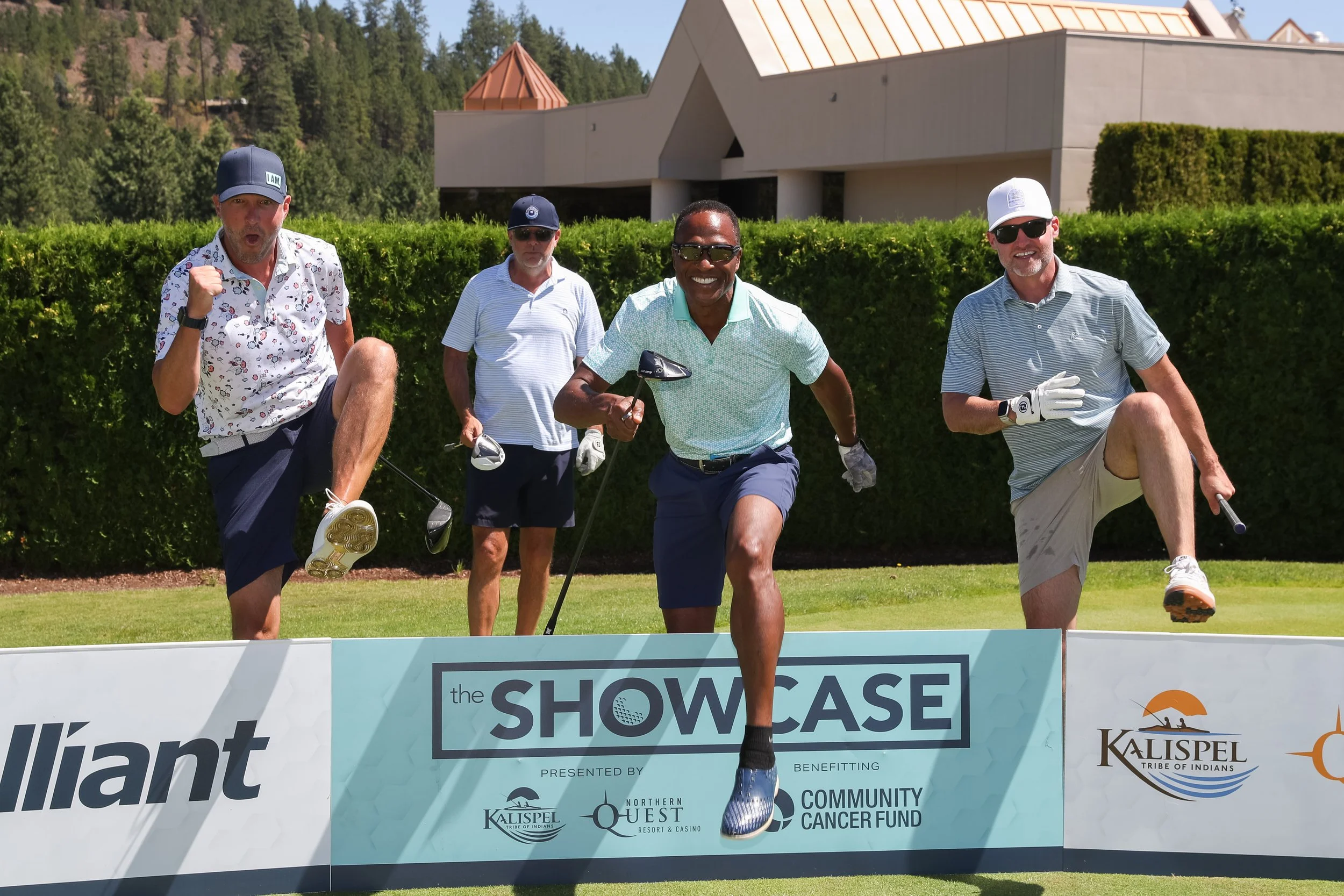 Five men participating in a golf event outdoors, jumping over a banner that reads 'the SHOWCASE' and includes logos for Kalispel Tribe of Indians, Northern Quest Resort & Casino, and Community Cancer Fund. The background features a building with a mo