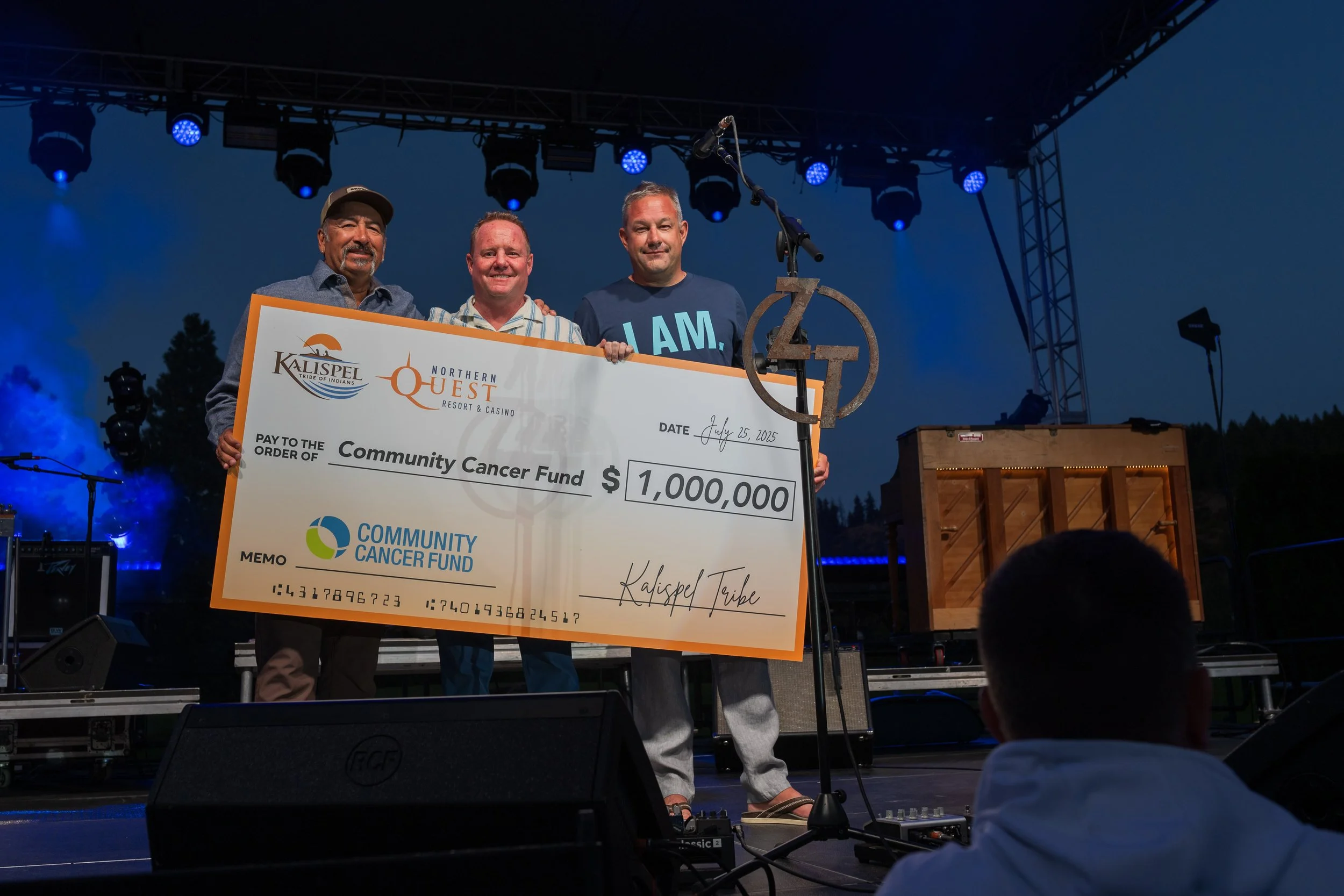 Three men on a stage holding a large check for $1,000,000 made out to the Community Cancer Fund. The check is from Kalispel Tribe and Northern Quest Resort & Casino, dated July 15, 2023. The men are smiling, with blue stage lights and musical equipme