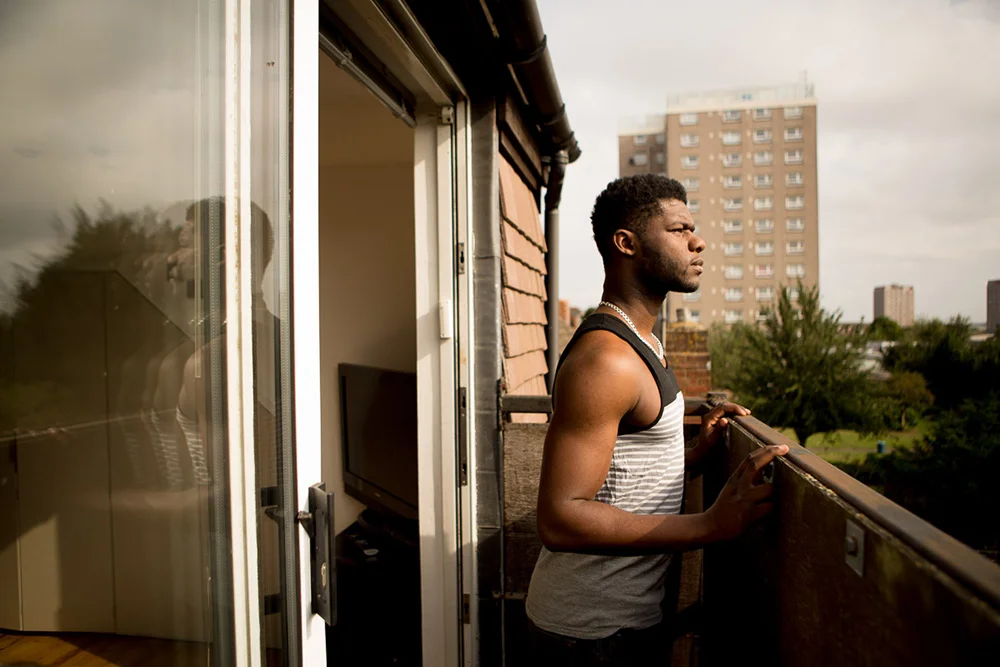 Junior looking out from the balcony of his house, Erith, 2015 Junior, always looking to secure his future, bought a house with his girlfriend Munjeet when they were both 23 years old; they lived on tins of tuna for 3 months to get the deposit together. Growing up in a council estate, he had never imagined being able to own his own property. Although his house is almost an hour on the train from Hackney, Junior is happy to be surrounded by more trees than concrete. He says his heart will always be in Hackney, he's even on the board of the Shoreditch Trust overseeing community projects in the area, but because of the gentrification, Hackney is not for people like him anymore. 