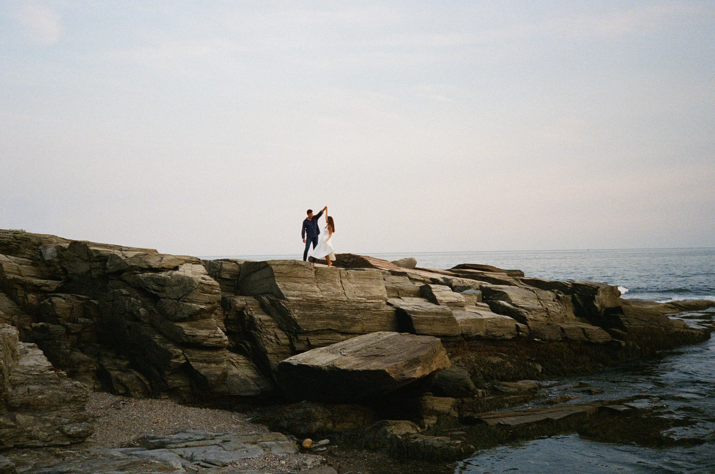 A couple dancing on rocky shoreline by the ocean under an overcast sky.