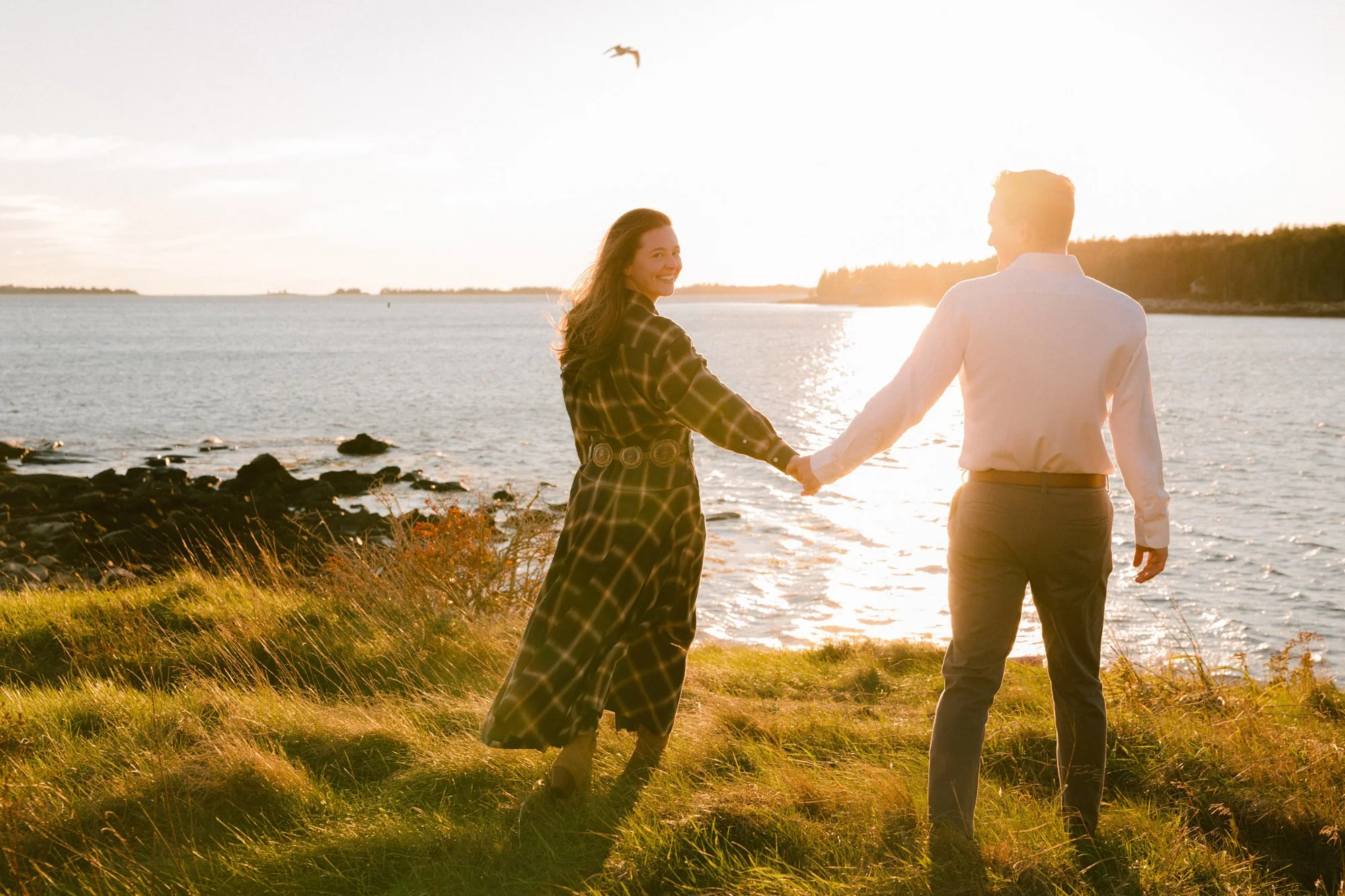 A couple holding hands and walking along a grassy shoreline during sunset, with water and distant land in the background and a seagull flying overhead.