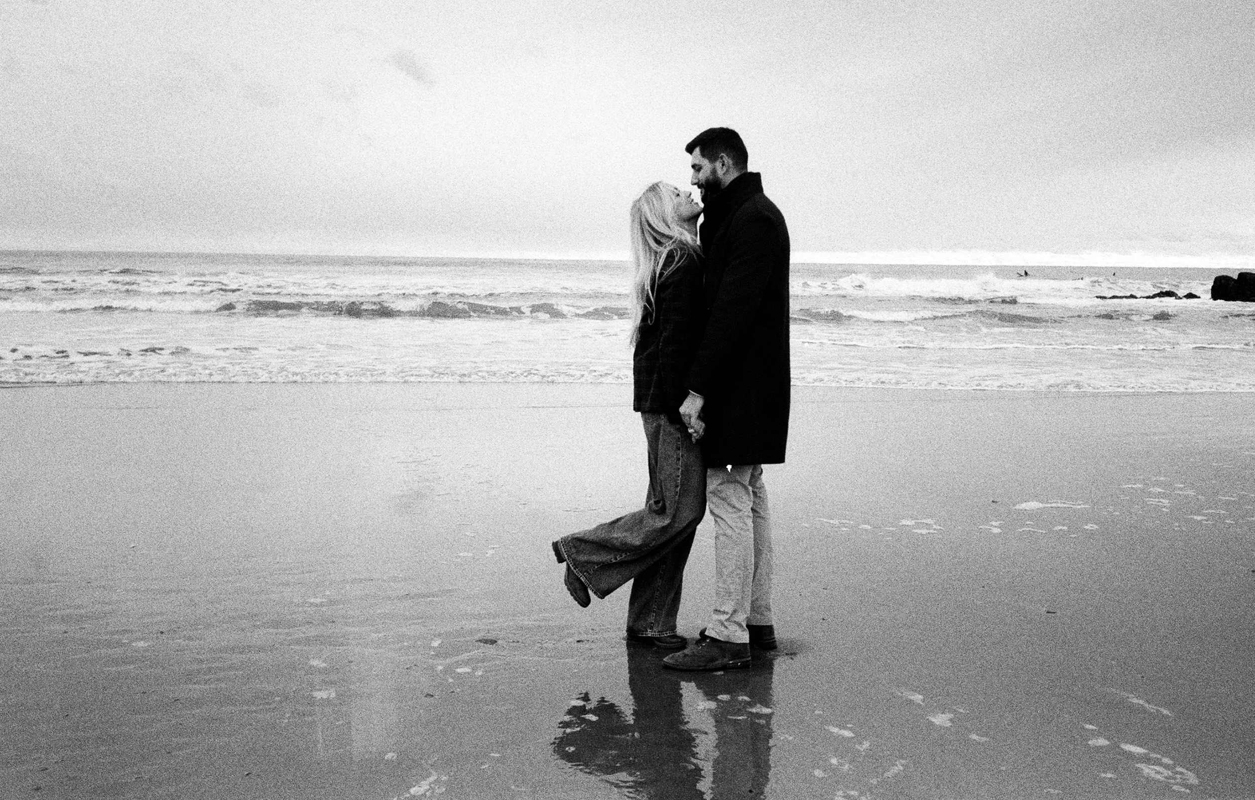 A black-and-white photo of a couple standing on the beach, gazing into each other's eyes, with the ocean and waves in the background.