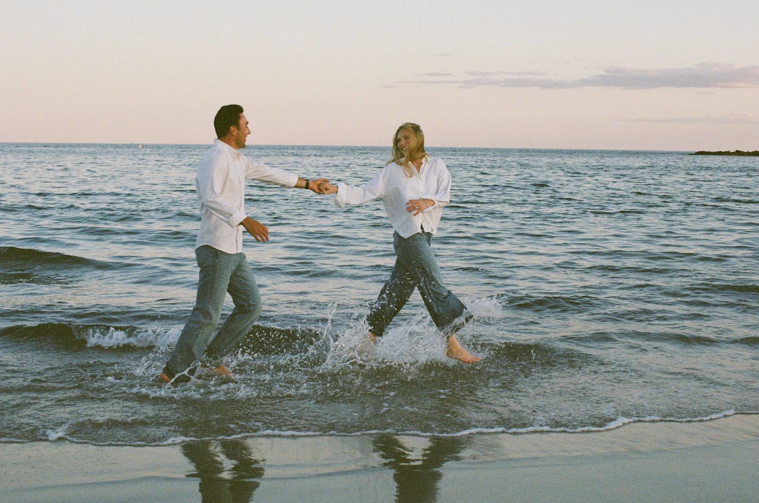 A 35 mm film photograph of a couple dressed in white shirts and pants holding hands while walking in the ocean water during sunset.